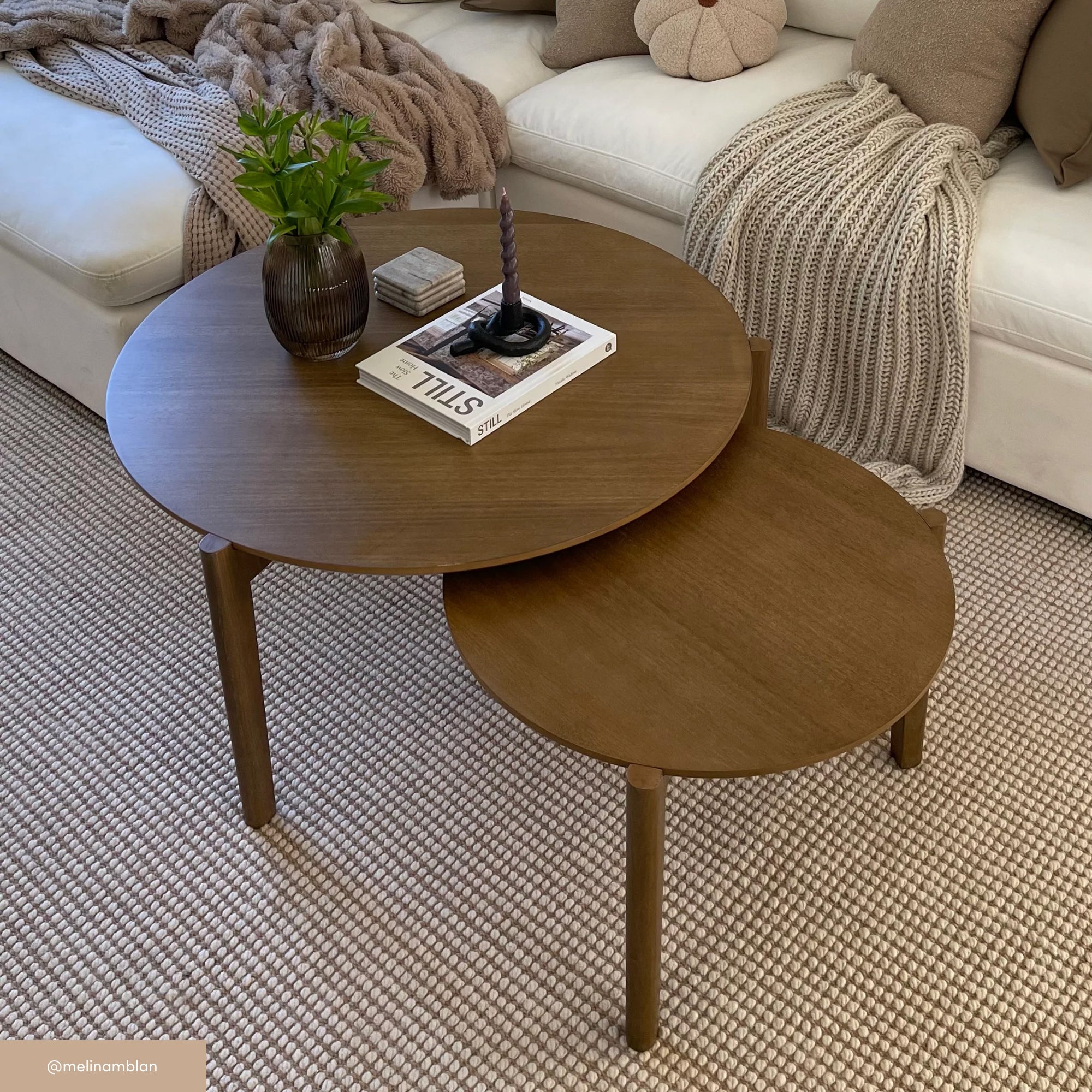 Two round wooden nesting coffee tables on a textured rug. The larger table holds a vase with green leaves, a candle, and a stack of books. In the background, a beige sofa with cozy pillows and a chunky knit throw.