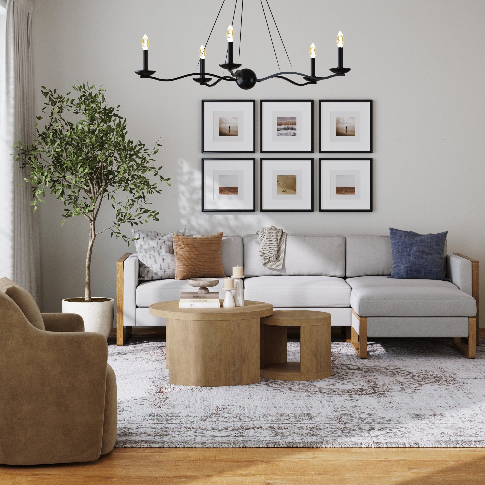 A modern living room with a light gray sectional sofa, a potted tree, a beige armchair, and the Nathan James Round Wood Nesting Coffee Table Set in light brown. Six framed art pieces decorate the wall beneath a black chandelier.