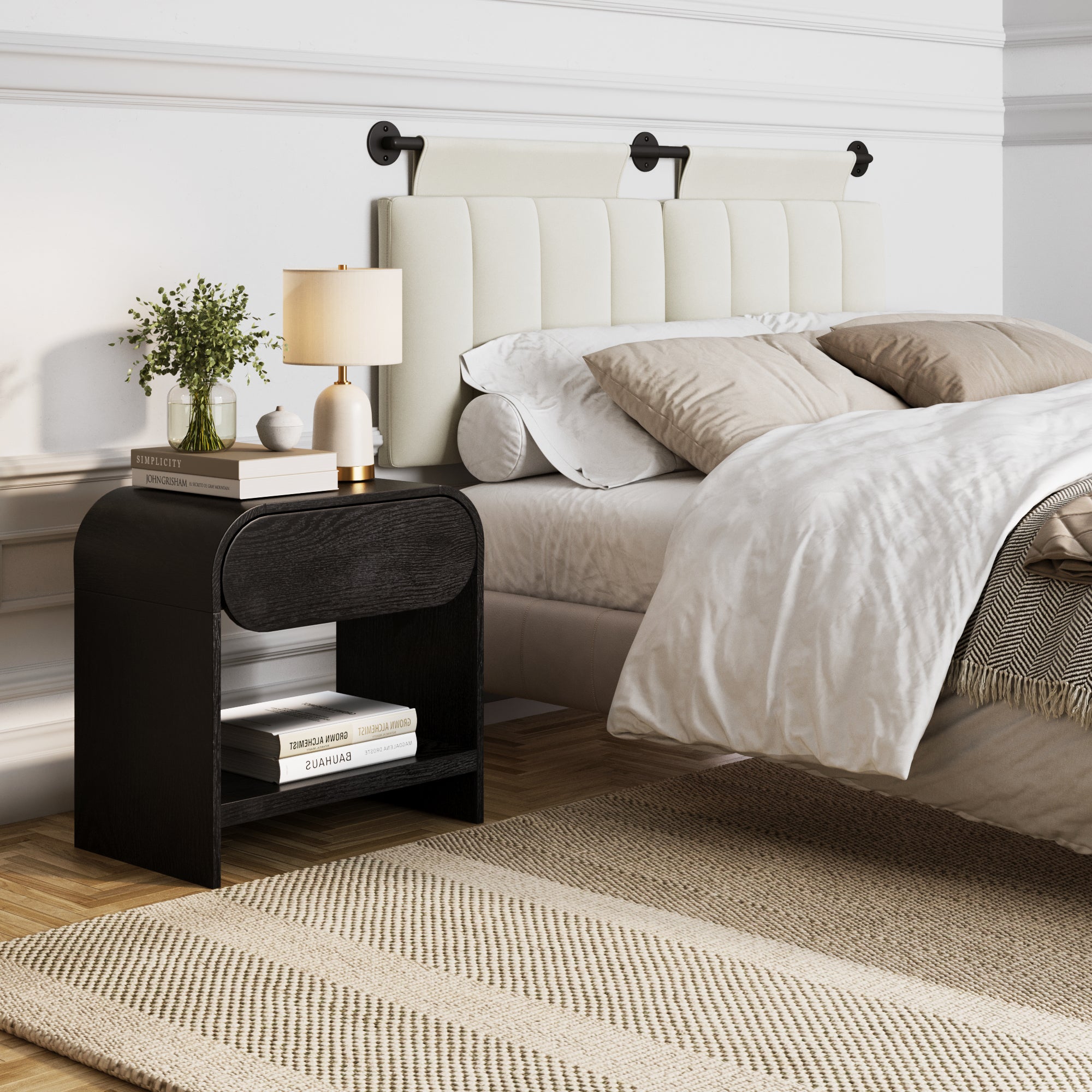 A modern bedroom with a bed featuring a padded white headboard, beige bedding, and a black nightstand holding books, a gold lamp, and a vase with greenery, set on a woven rug over a wooden floor.
