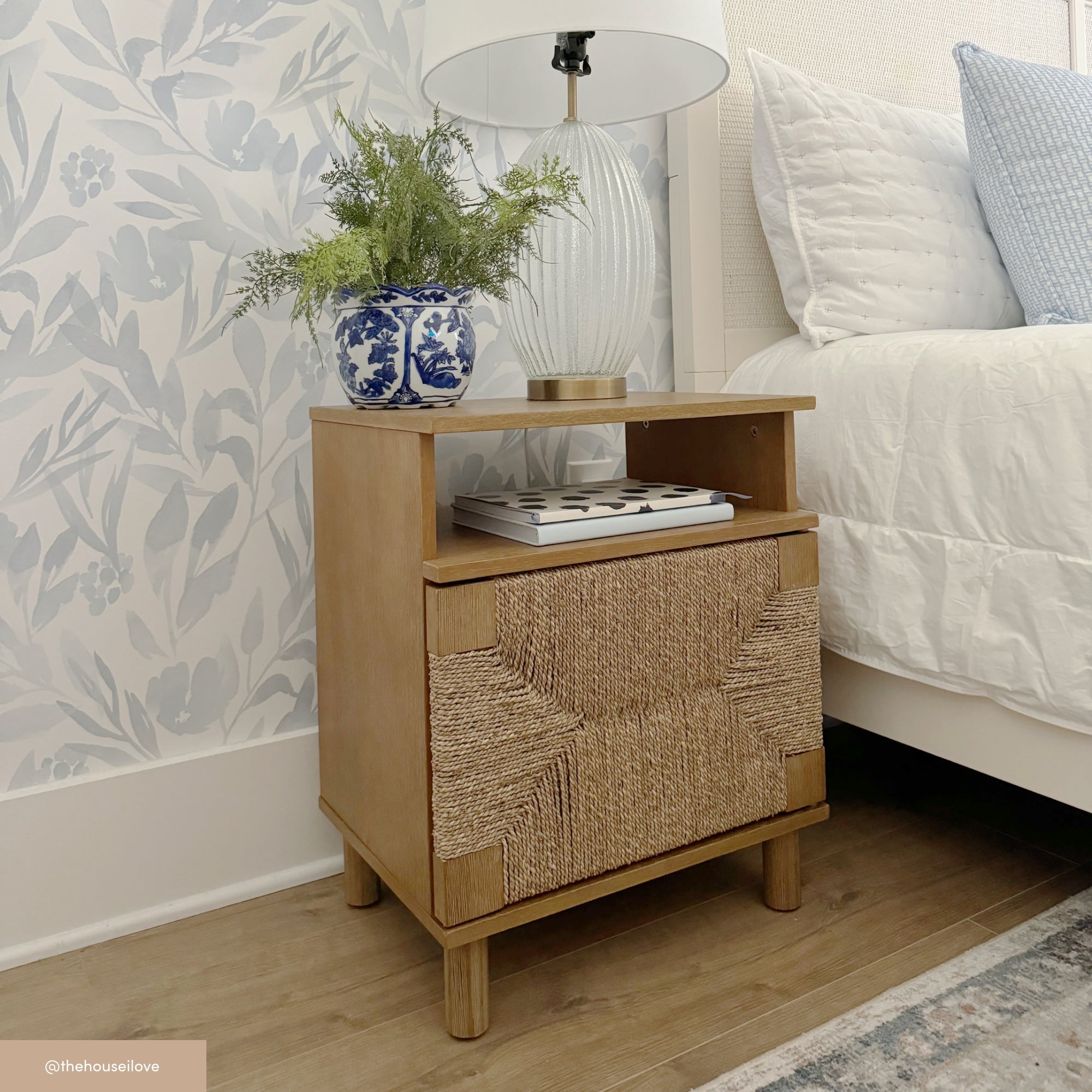 A wooden nightstand with a woven front panel holds a white lamp with a ribbed glass base, a blue and white planter with green foliage, and two books. The stand is beside a bed with white and blue bedding and floral wallpaper.