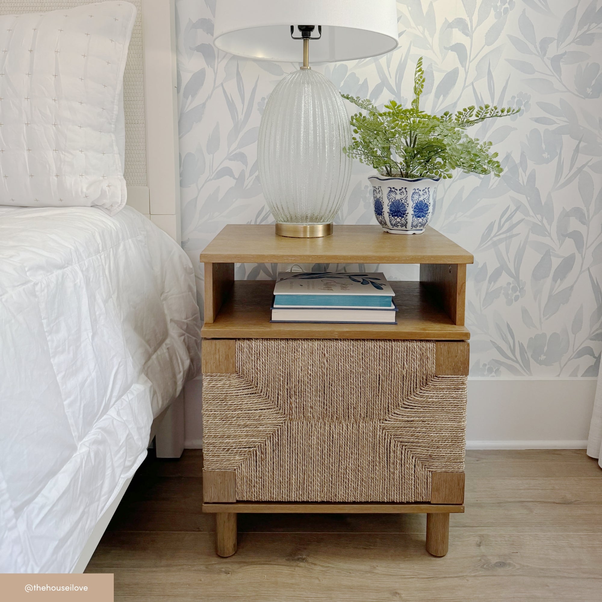 A wooden nightstand with a woven front sits beside a bed. On top, there is a white textured lamp with a gold base, a blue and white planter with greenery, and a neutral wallpaper with leafy designs in the background.