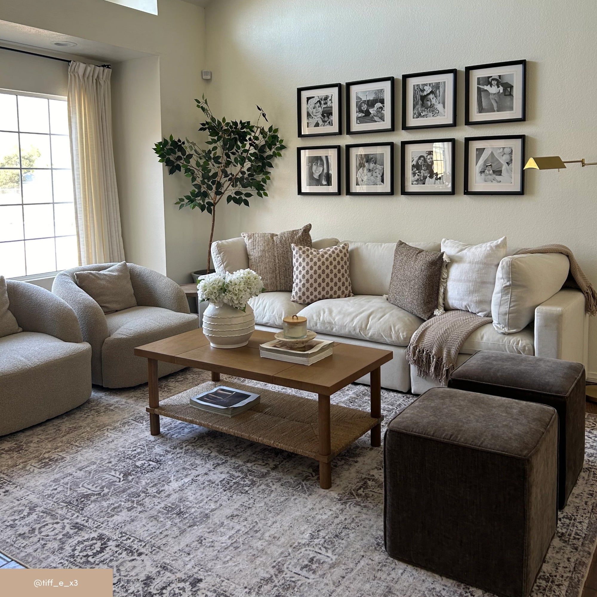 A cozy living room with a white sofa, patterned pillows, two brown ottomans, two gray armchairs, a wooden coffee table, a rug, a potted plant, and a gallery wall with nine black-and-white photos. Sunlight streams in through a window.