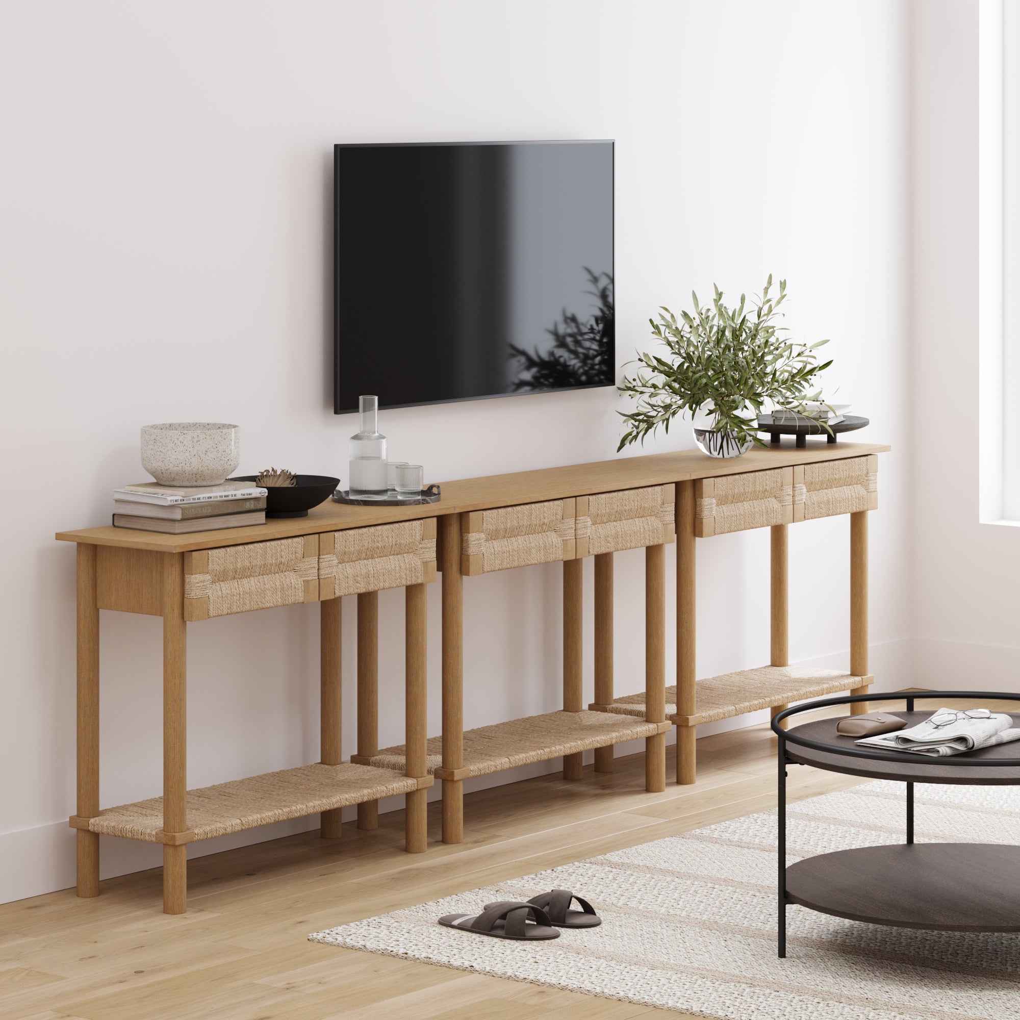 A modern living room showcases the Nathan James Boho Seagrass Console Tables (Set of 3) beneath a wall-mounted TV, styled with decor like plants and bowls. A round coffee table adds both style and function in the foreground.