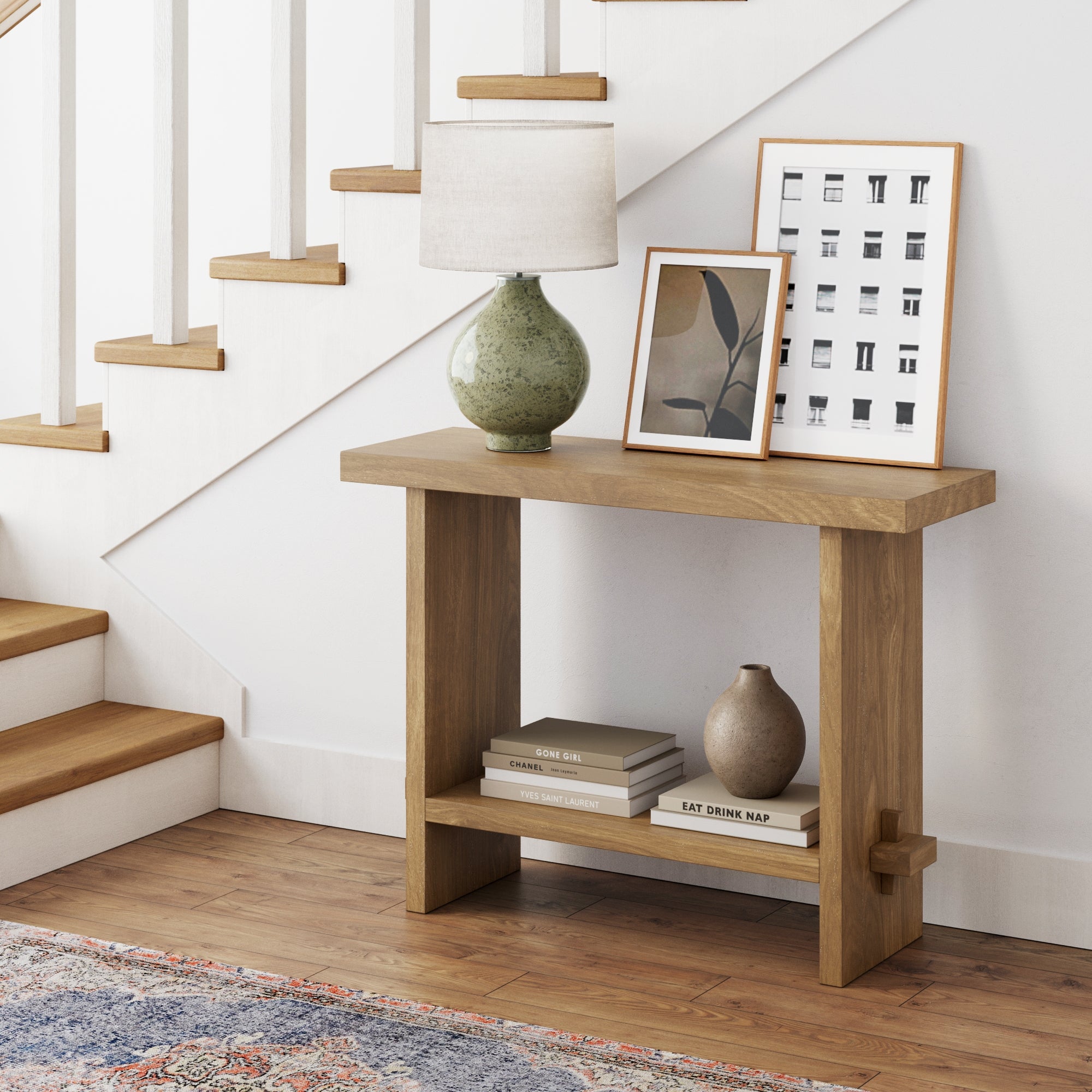 The Nathan James Wood Console Table in Light Brown sits under a staircase, displaying a green lamp, framed artwork, and a vase. Another vase and two books rest on the lower shelf. A cozy rug and warm wood floor complete the scene.