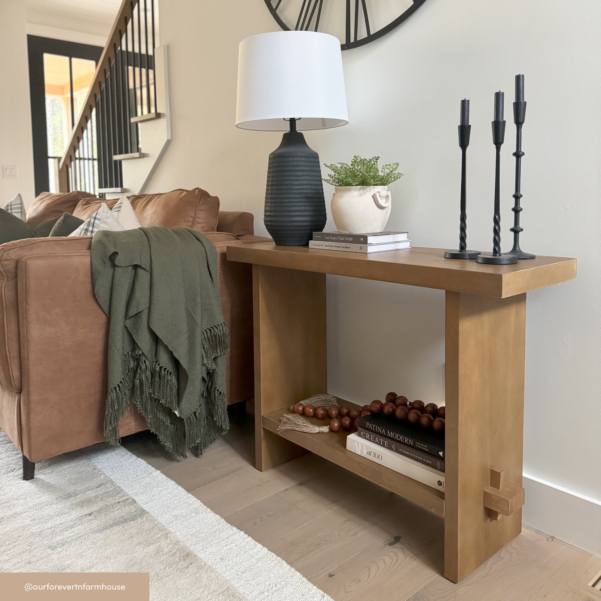 A wooden console table beside a brown leather sofa holds a black lamp, a small plant, books, decorative beads, and two black candle holders. A green throw blankets the sofa; a black wall clock hangs above.