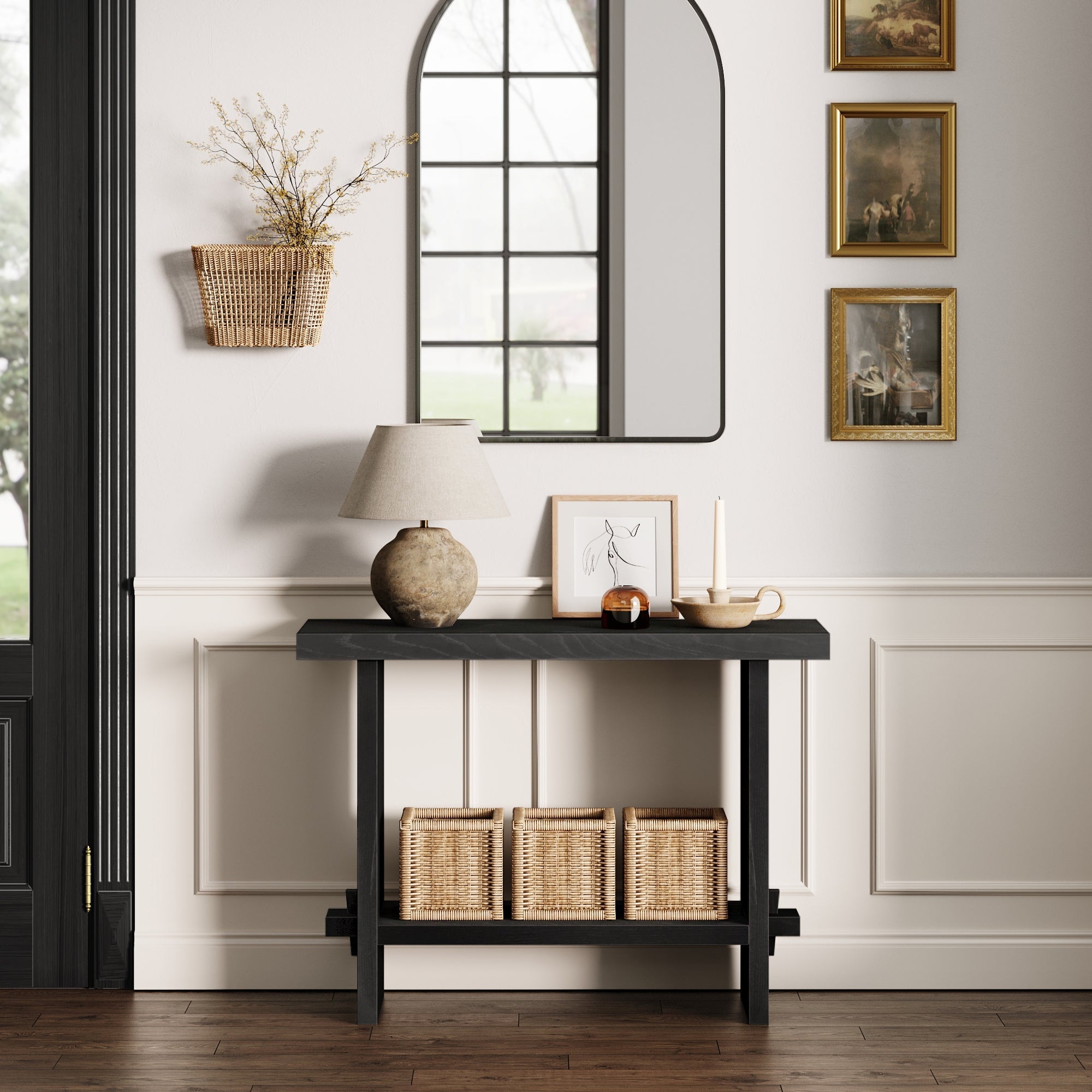 The Nathan James Wood Console Table in Black Oak sits against a white wainscoted wall, styled with a lamp, framed art, and candles. Three woven baskets are underneath, while a mirror and wicker basket hang above.