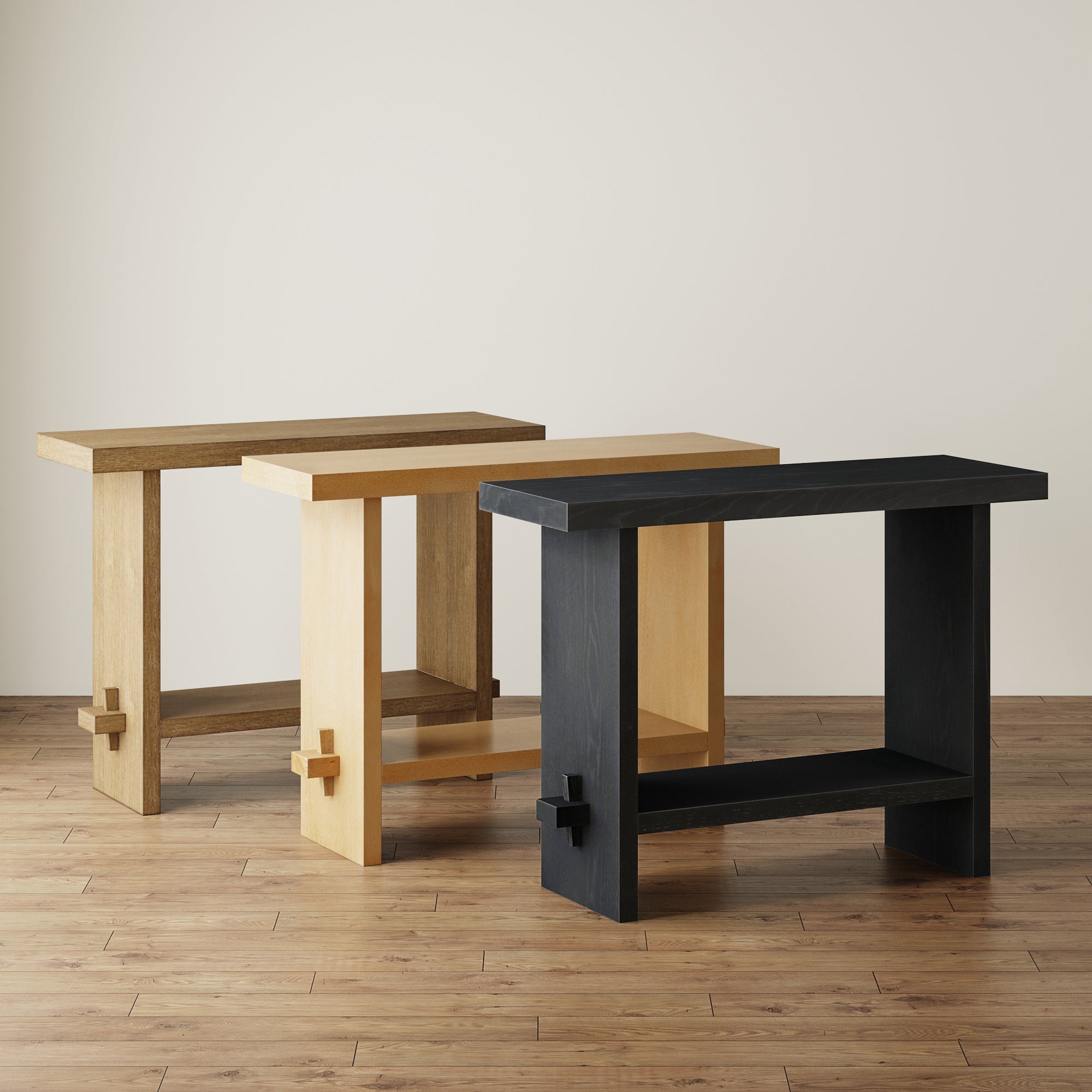 Three wooden console tables in different finishes—brown, light wood, and black—are arranged on a hardwood floor against a plain light wall. Each table features a modern, minimalist design with a cross detail on one leg.
