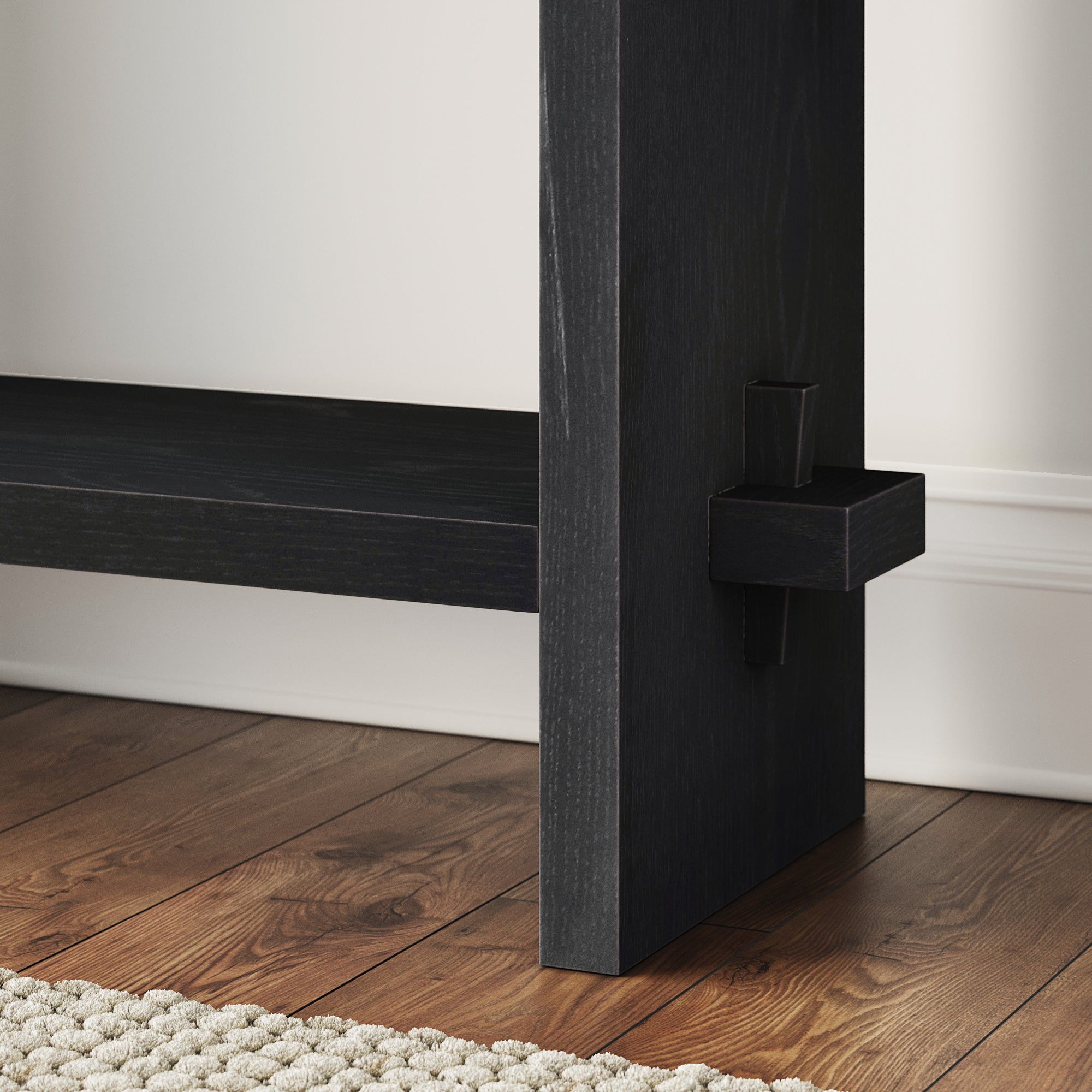 Close-up of a black wooden furniture leg on a hardwood floor next to a textured beige rug, with a white wall and baseboard in the background.