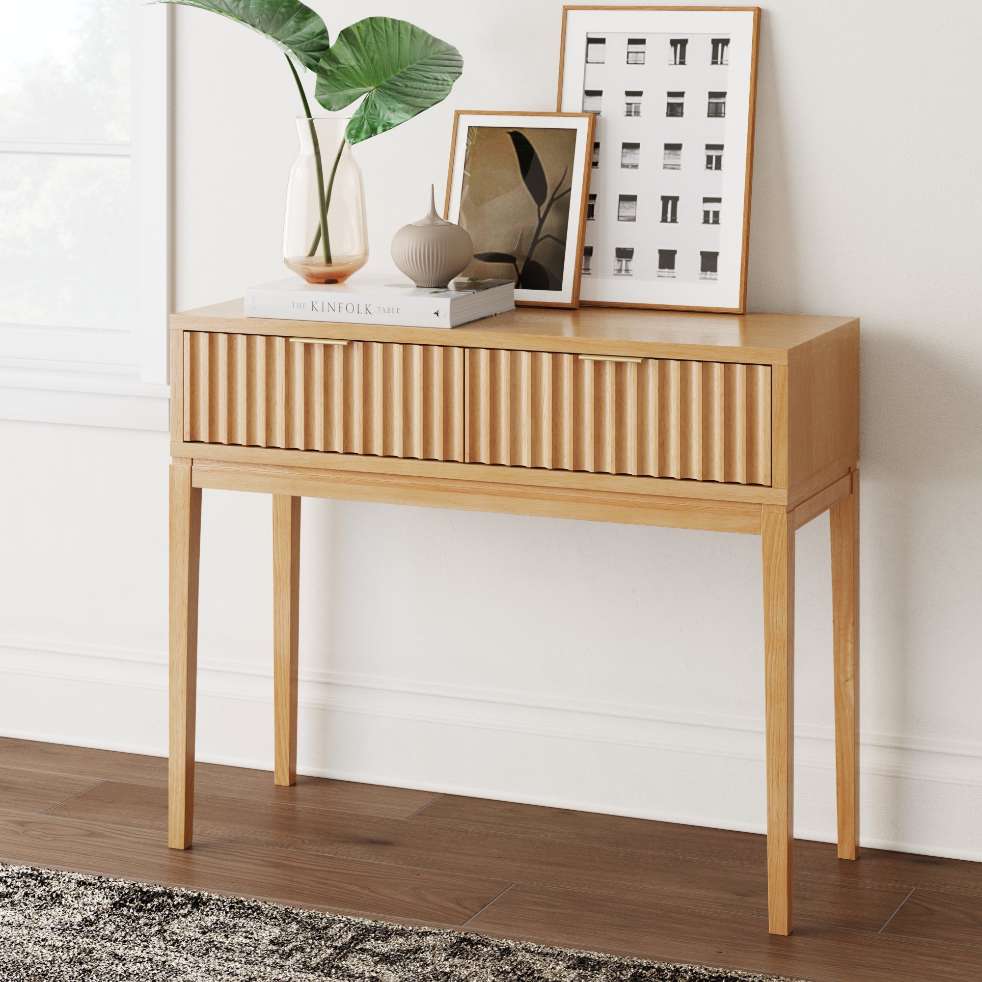 The Nathan James Mid-Century Fluted 2-Drawer Console Table stands against a white wall, topped with two framed art prints, green-leafed and beige vases, and a book. A window is partially visible on the left.