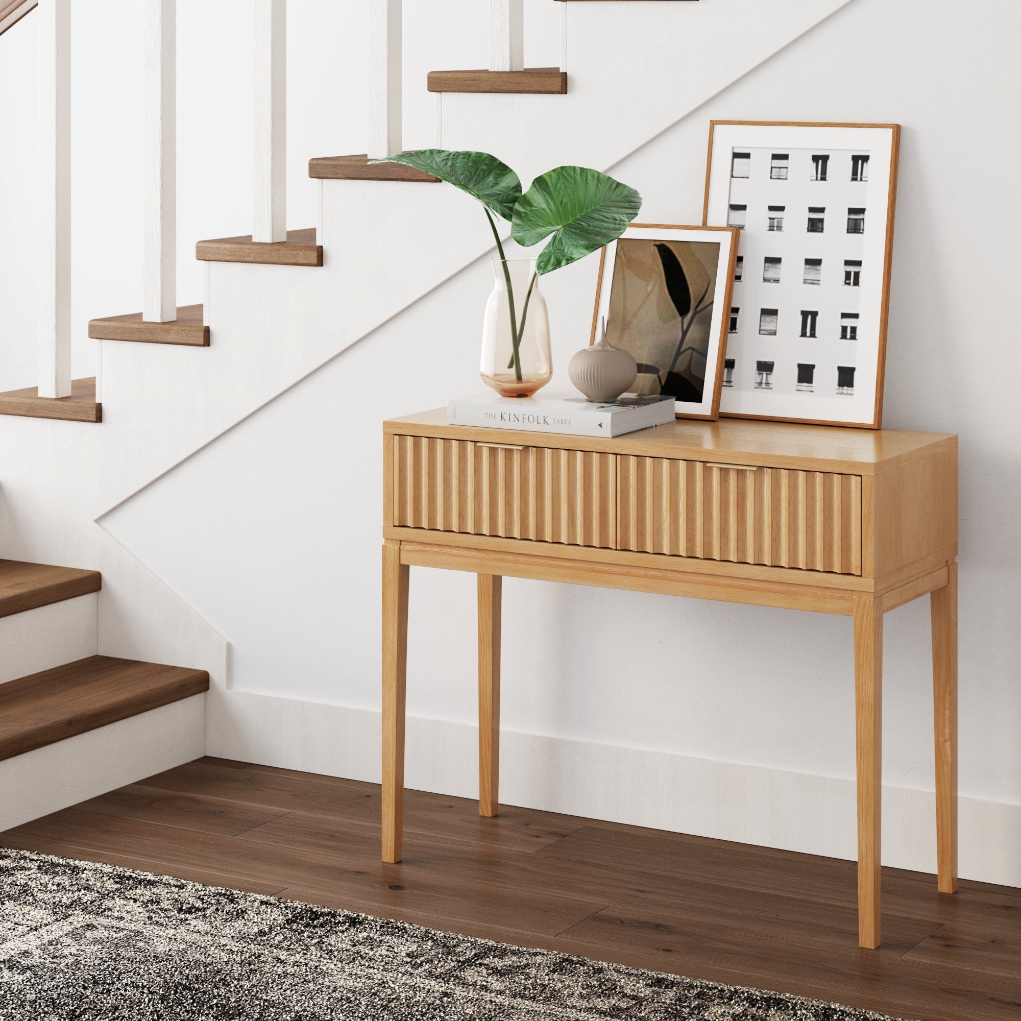 The Nathan James Mid-Century Fluted 2-Drawer Console Table stands by the staircase, displaying books, art prints, and a vase of greenery against a white wall, dark wood stairs, and a patterned rug.