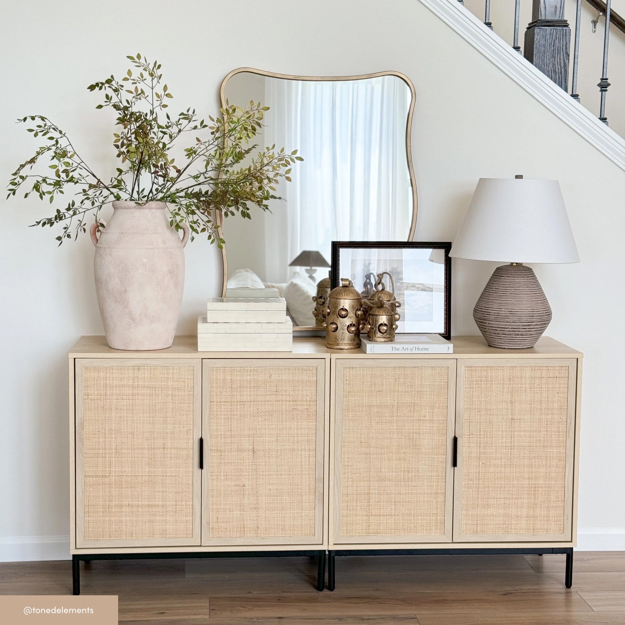 A light wood sideboard with four doors is decorated with a large vase of greenery, stacked books, gold decorative heads, a framed photo, and a textured table lamp. A mirror hangs above, reflecting a staircase.