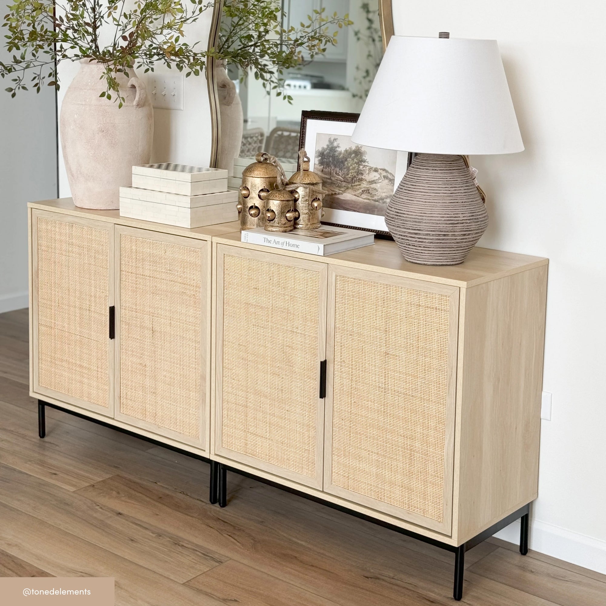 A light wood sideboard with rattan doors displays a striped ceramic lamp, two gold decorative jars, books, a framed picture, and tall vases with greenery, set against a white wall and large mirror.