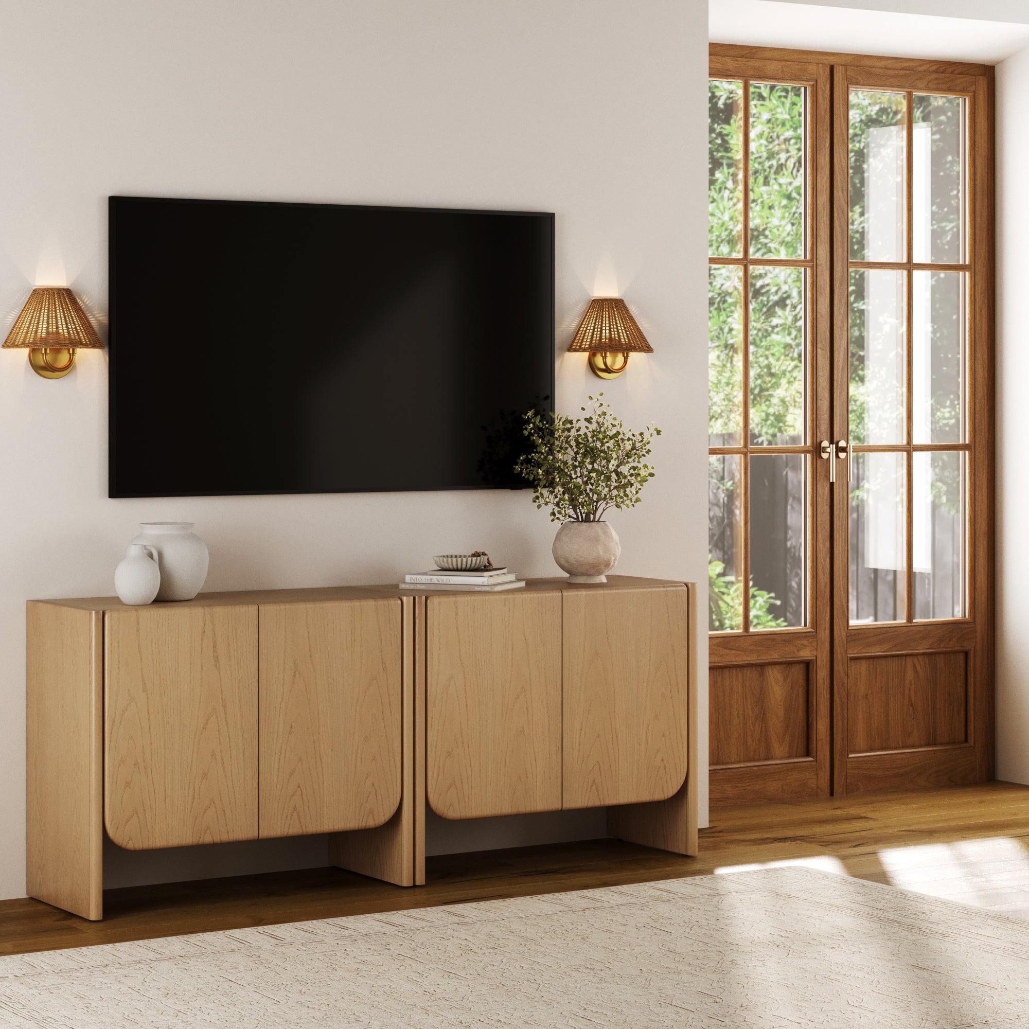 A modern living room featuring a set of two Nathan James Rounded Wood Accent Cabinets in Raw Oak used as a TV stand, with decorative vases, books, wall-mounted TV, sconces, and French doors allowing natural light.
