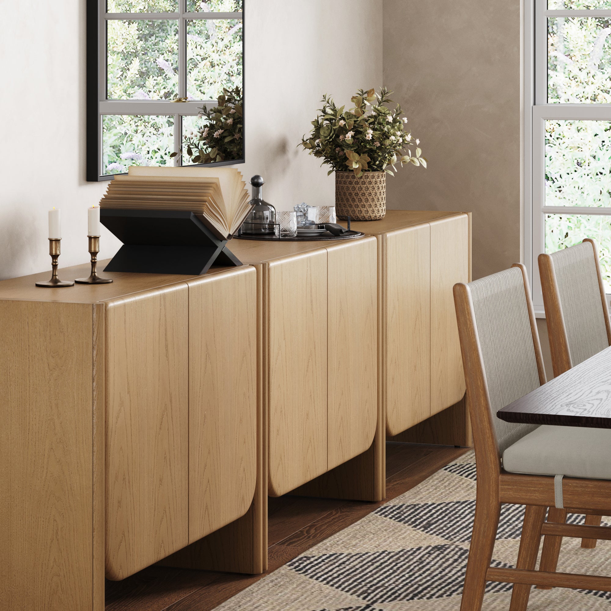 The Nathan James Rounded Wood Accent Cabinet or TV Stand in Raw Oak (Set of 3) with curved edges displays a potted plant, two candlesticks, an open book on a stand, and glassware beside a dining table near sunlit windows.