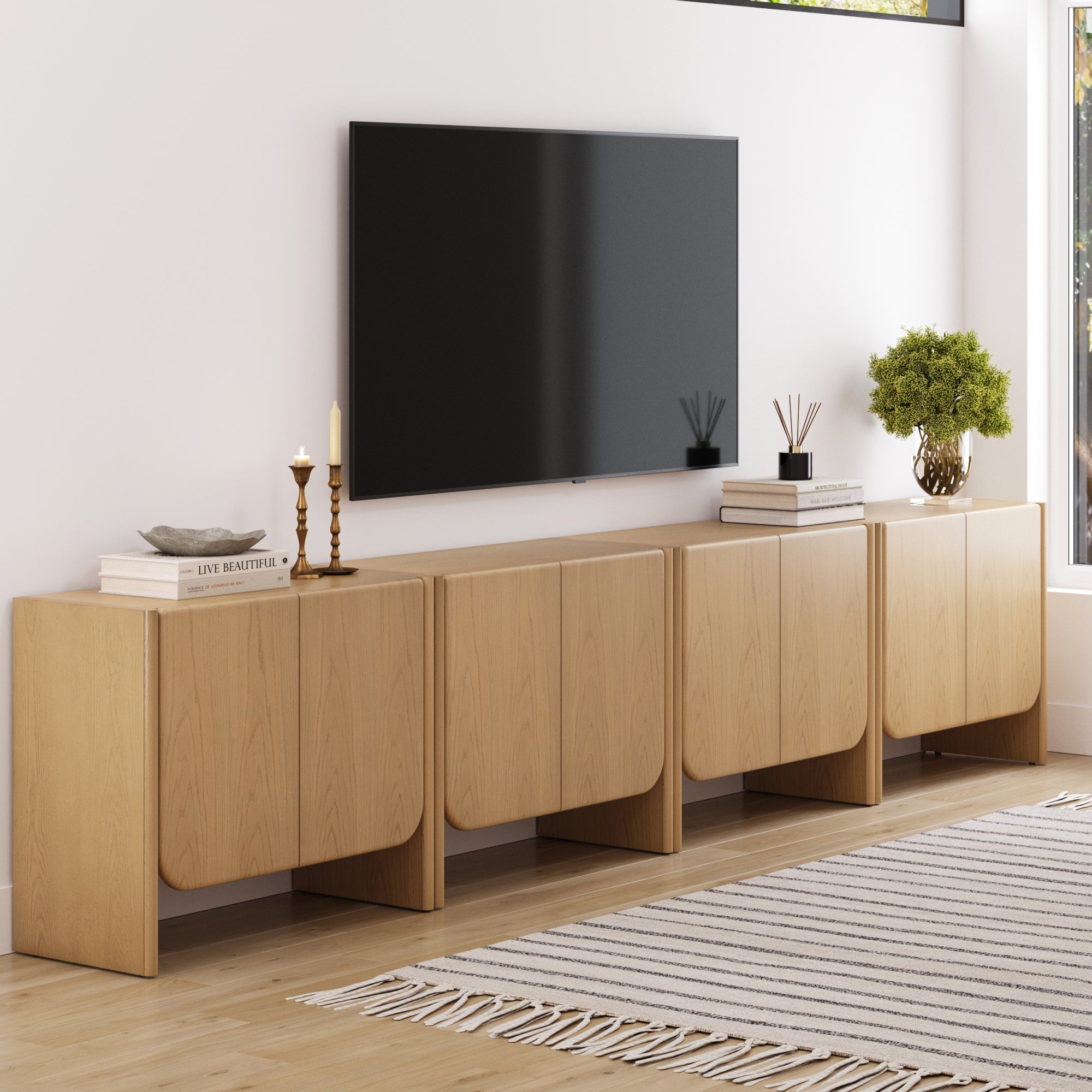 A modern living room features a large wall-mounted TV above the Nathan James Rounded Wood Accent Cabinet or Large Credenza Raw Oak (Set of 4), decorated with books, candles, a plant, and a reed diffuser. A striped rug covers the wooden floor.