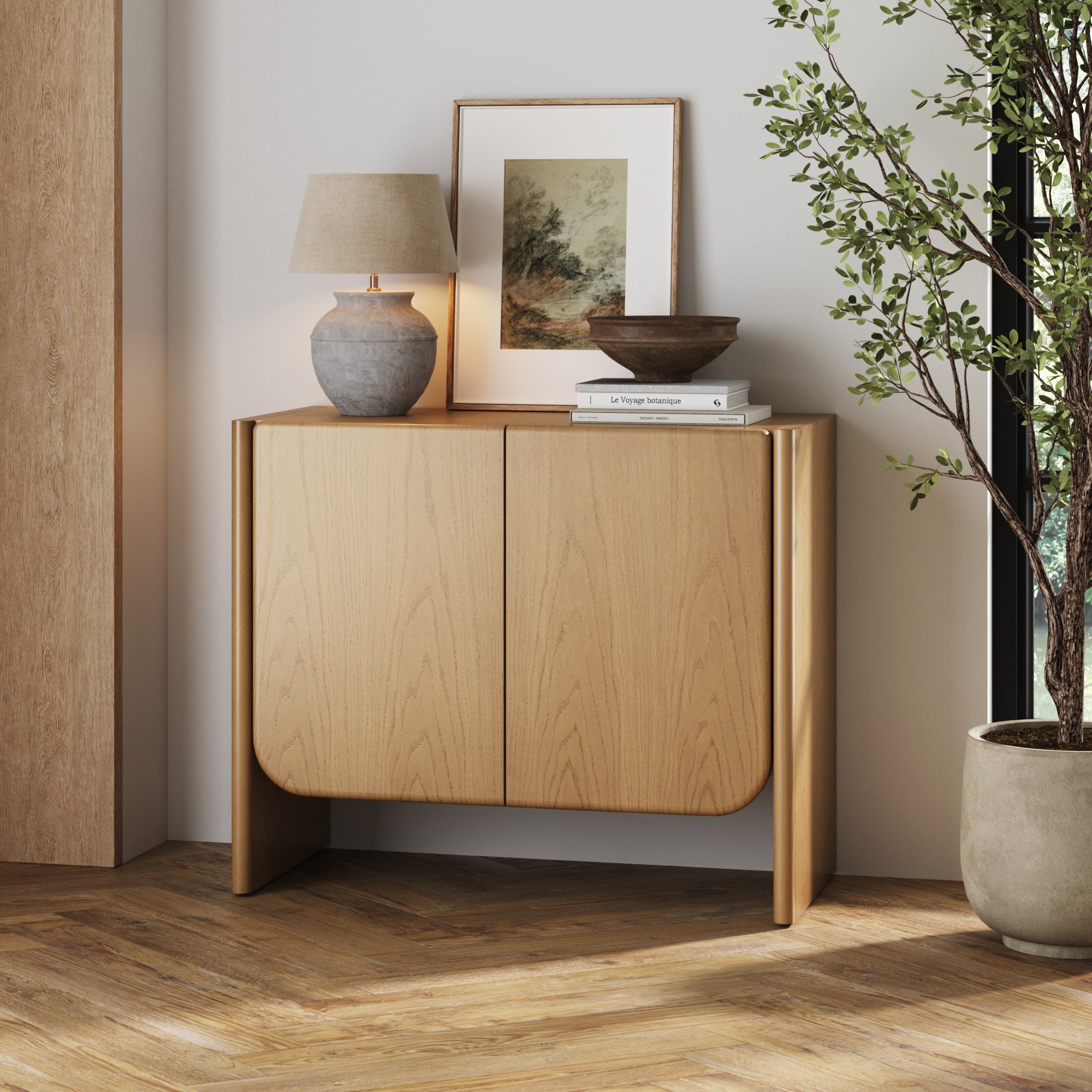 The Nathan James Mid Century Modern Rounded Wood Accent Cabinet in raw oak, featuring two doors, stands against a chevron wood floor. On top rest a ceramic lamp, framed picture, books, and bowl; a potted plant stands beside it.