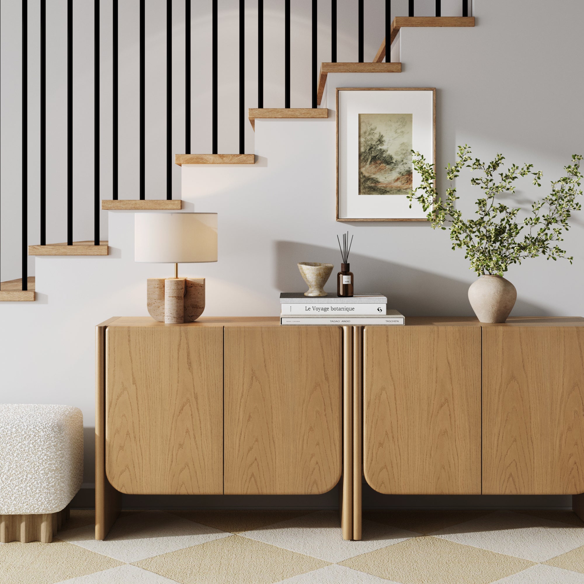A modern interior showcases the Nathan James Mid Century Modern Rounded Wood Accent Cabinet in Raw Oak, paired with a table lamp, books, decor, and a green-branch vase beneath open wooden stairs with black railings in a minimalist style.