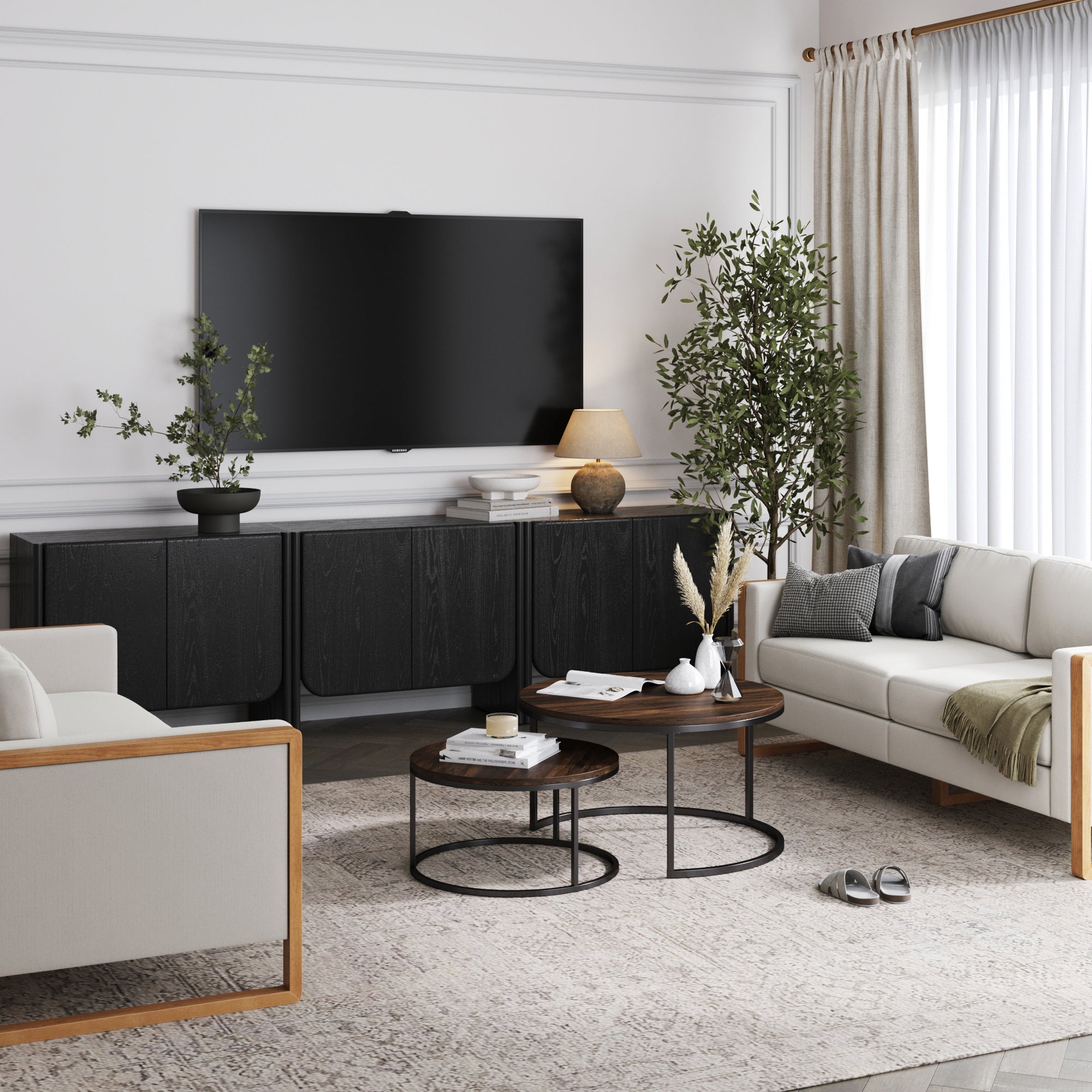A modern living room with a wall-mounted TV, Nathan James Rounded Wood Curio Cabinet or TV Stand in Black Oak (Set of 3), two round coffee tables, beige sofa, armchair, indoor plants, lamp, books, and a large window with sheer curtains.