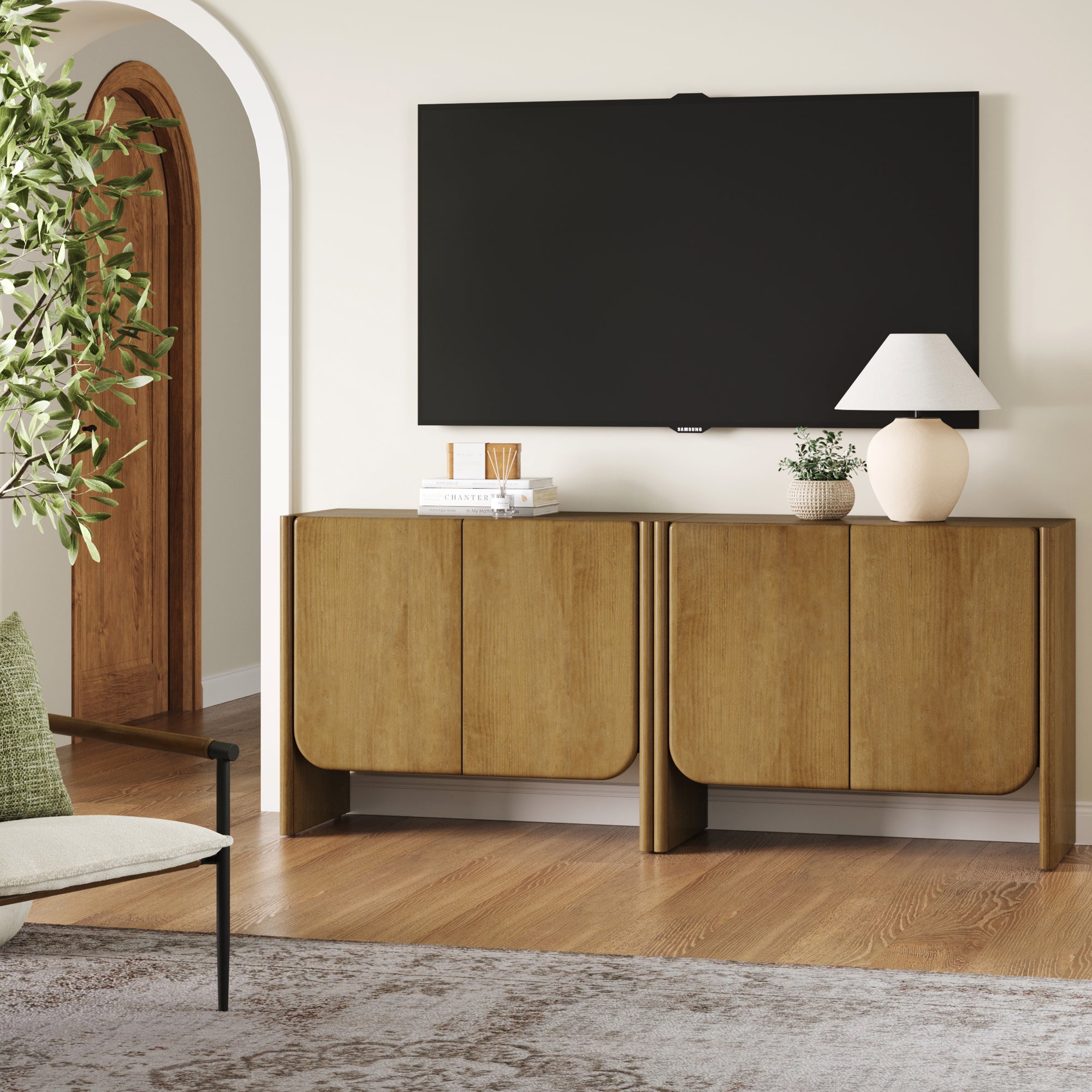 A modern living room with a wall-mounted TV above Nathan James Arched Wood Accent Cabinet or TV Stand Light Brown (Set of 2), a white table lamp, decor, a potted plant, an arched wooden door, a chair with green cushion, and a light rug.