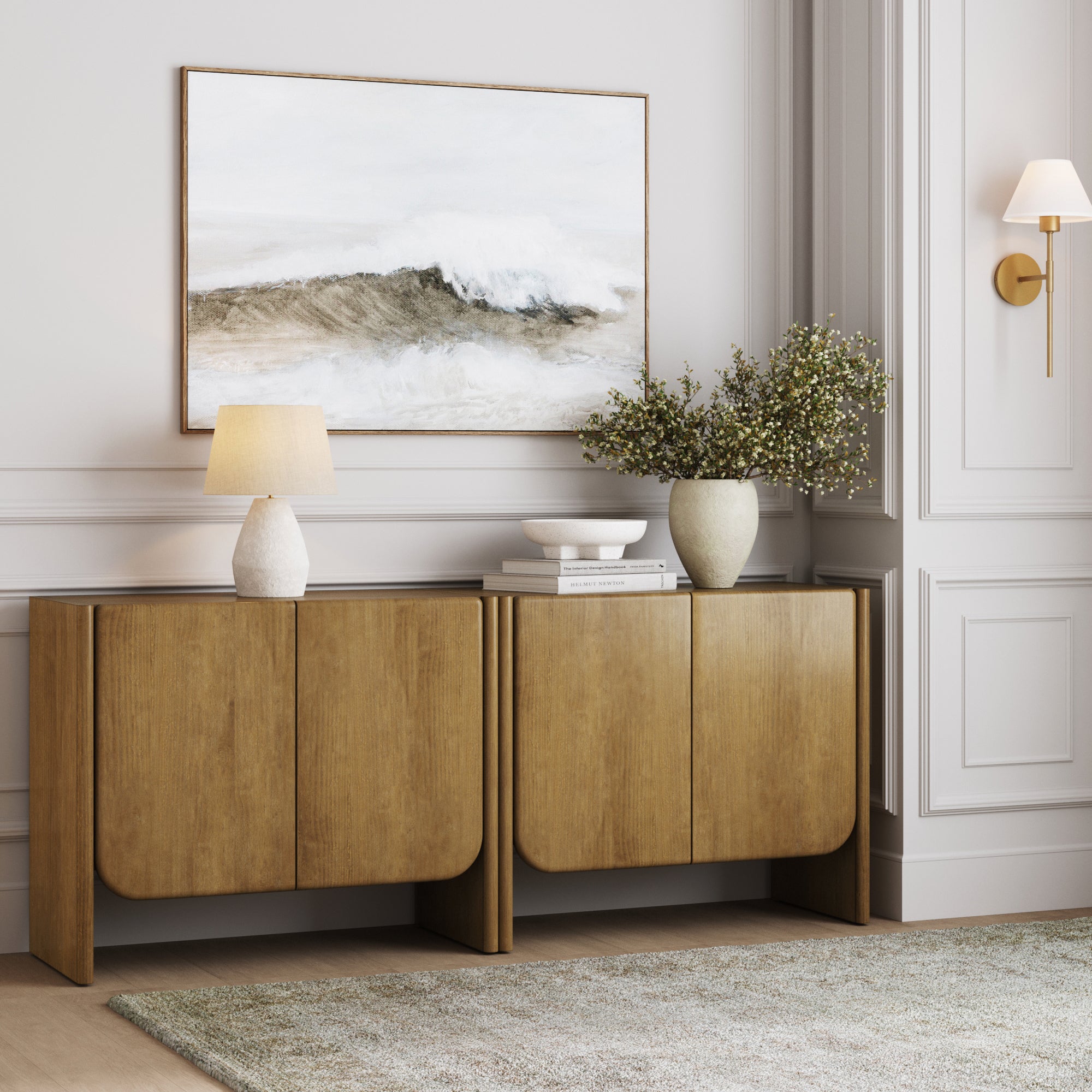 The Nathan James Arched Wood Accent Cabinets in light brown (set of 2) feature rounded edges and display a lamp, books, white bowl, and vase with greenery. A framed ocean wave painting hangs above on a light gray wall with molding and sconce.
