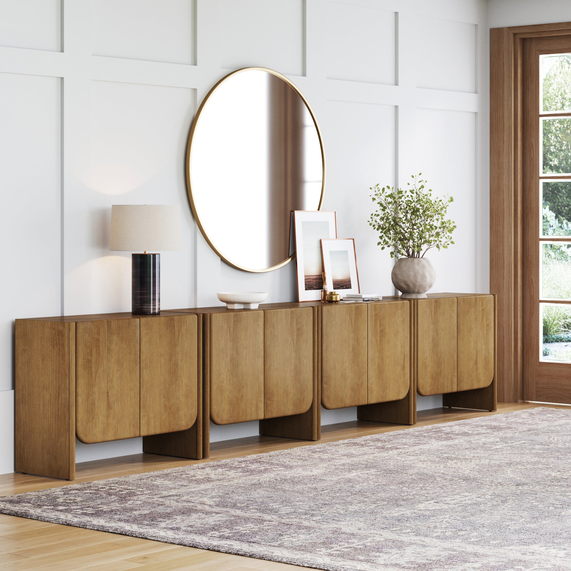 A modern living room features the Nathan James Arched Wood Accent Cabinet or Large Credenza Light Brown (Set of 4), a round mirror above, table lamp, framed photos, vase with greenery, light walls, large window, adjustable shelf, and patterned rug.
