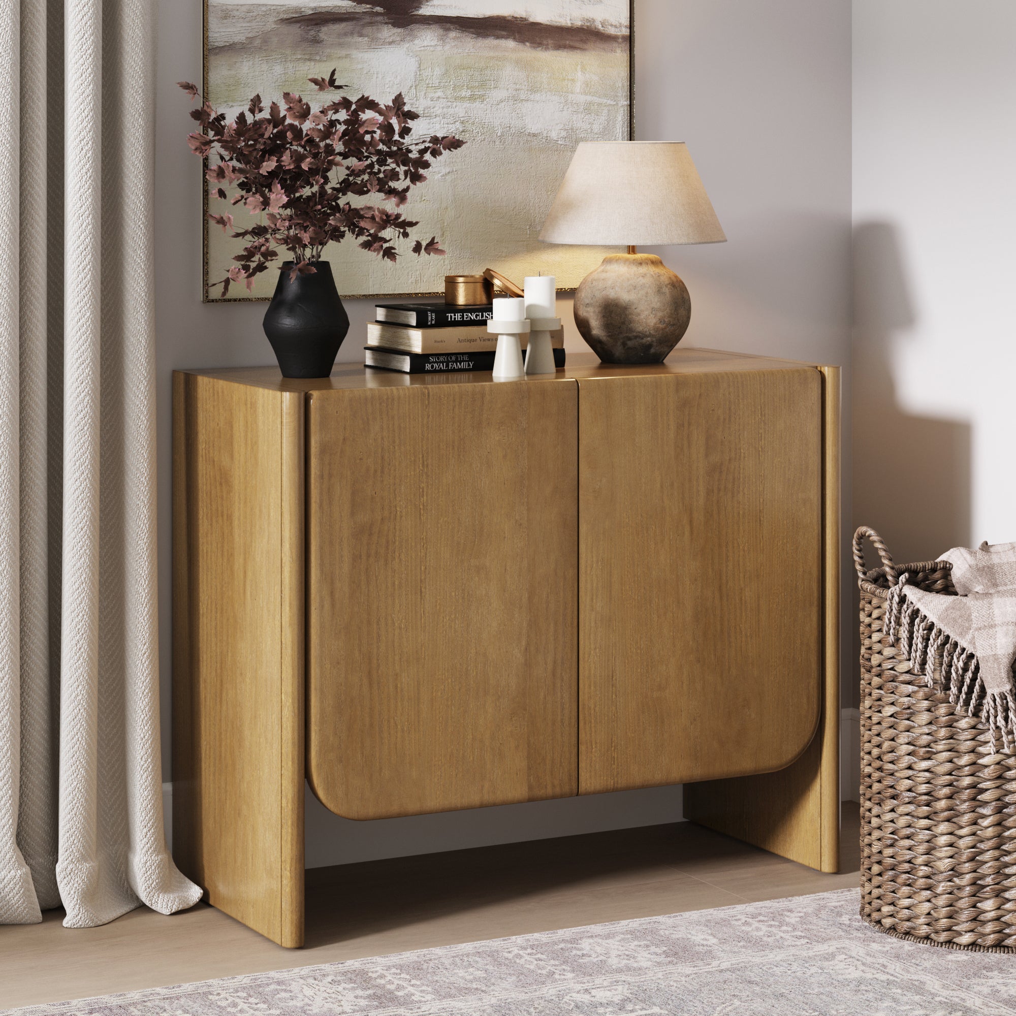 The Nathan James Mid Century Modern Arch Storage Cabinet in light brown stands against a wall, topped with books, a vase of dark foliage, two candles, and a lamp. Above hangs a large framed painting; nearby on the rug sits a woven basket.