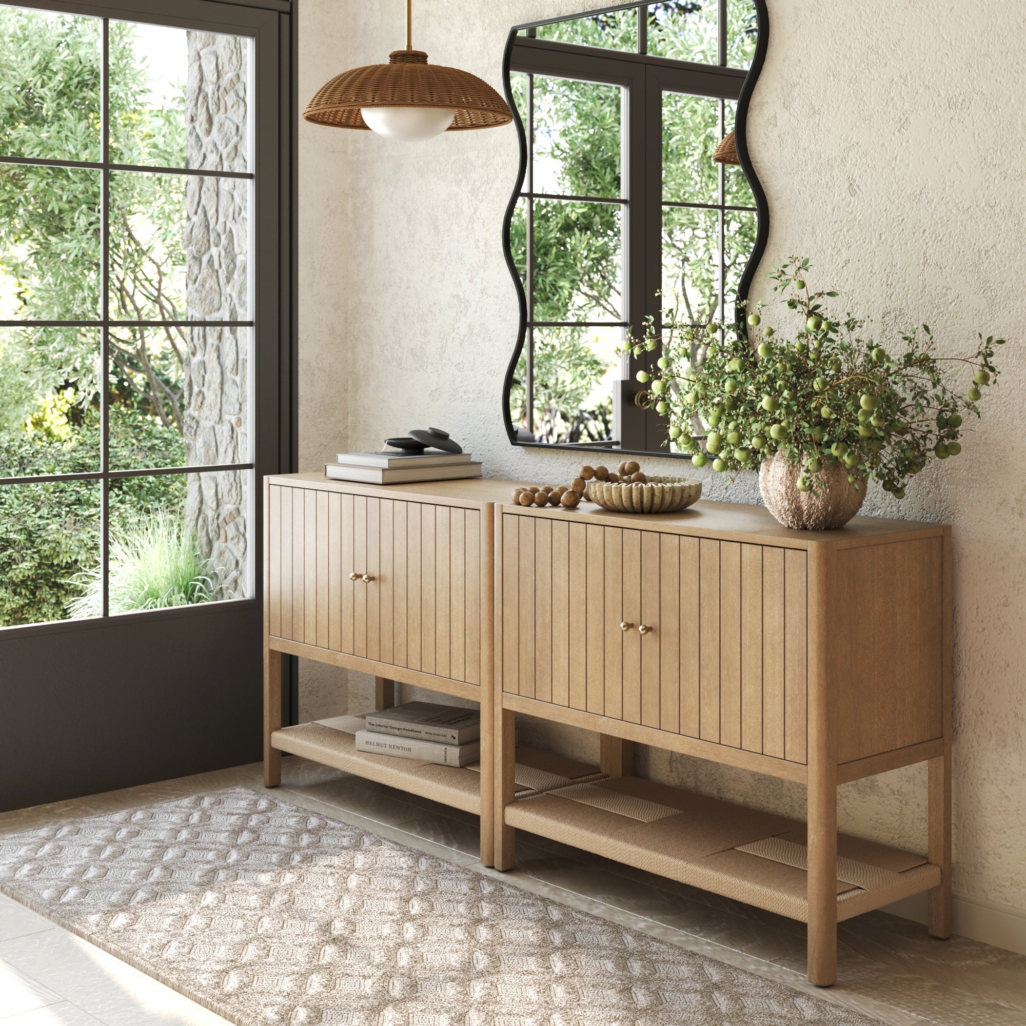 The Nathan James Grooved Door TV Stand or Storage Cabinet (Set of 2) in light brown stands against a cream wall, styled with greenery, a bowl, and tray. Above hangs a large wavy mirror with sunlight streaming through nearby glass-paneled doors.