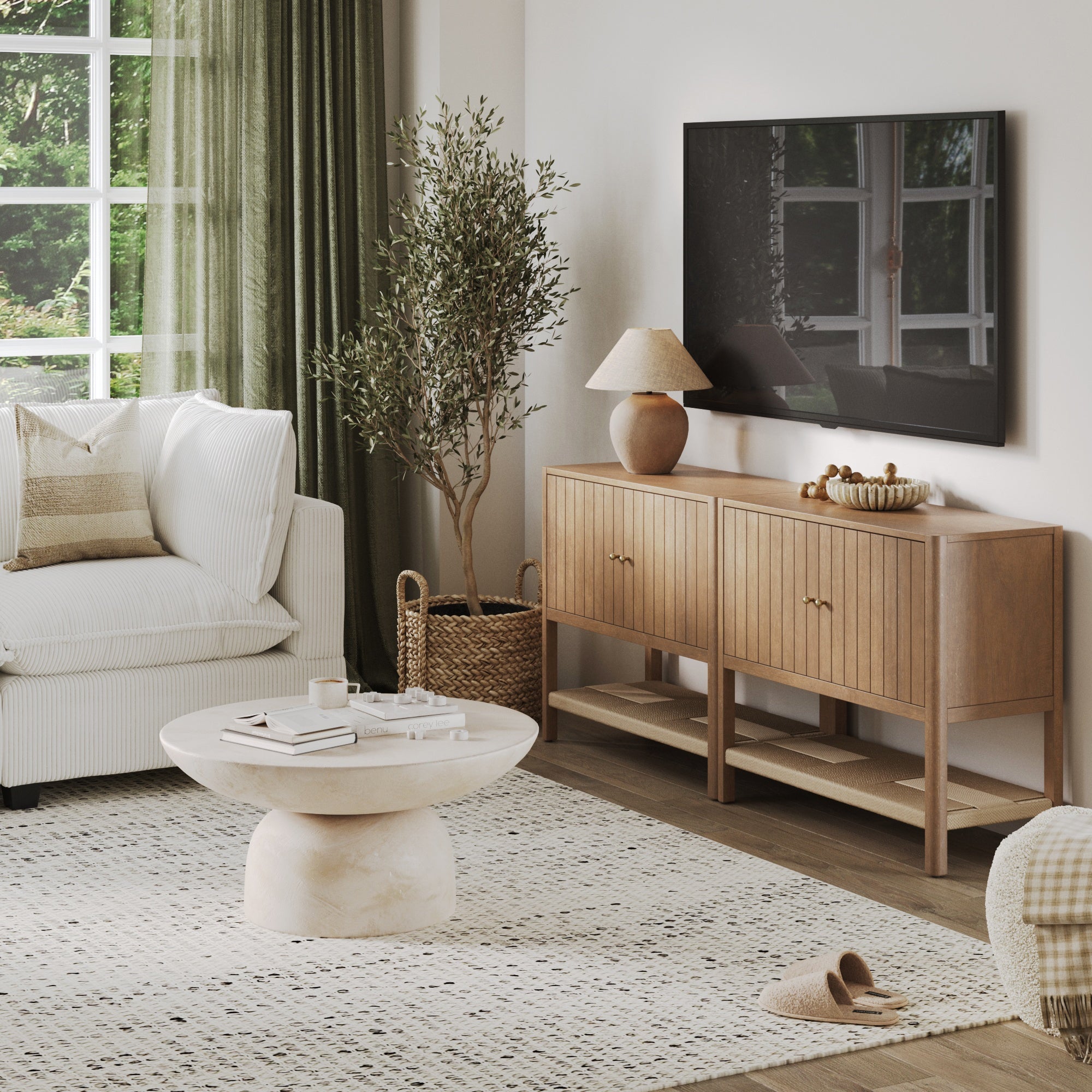 A modern living room featuring a white sofa, round marble coffee table, Nathan James Grooved Door TV Stand or Storage Cabinet Light Brown (Set of 2), table lamp, potted plant, wall-mounted TV, large window with green curtains, and a patterned rug on hardwood floors.