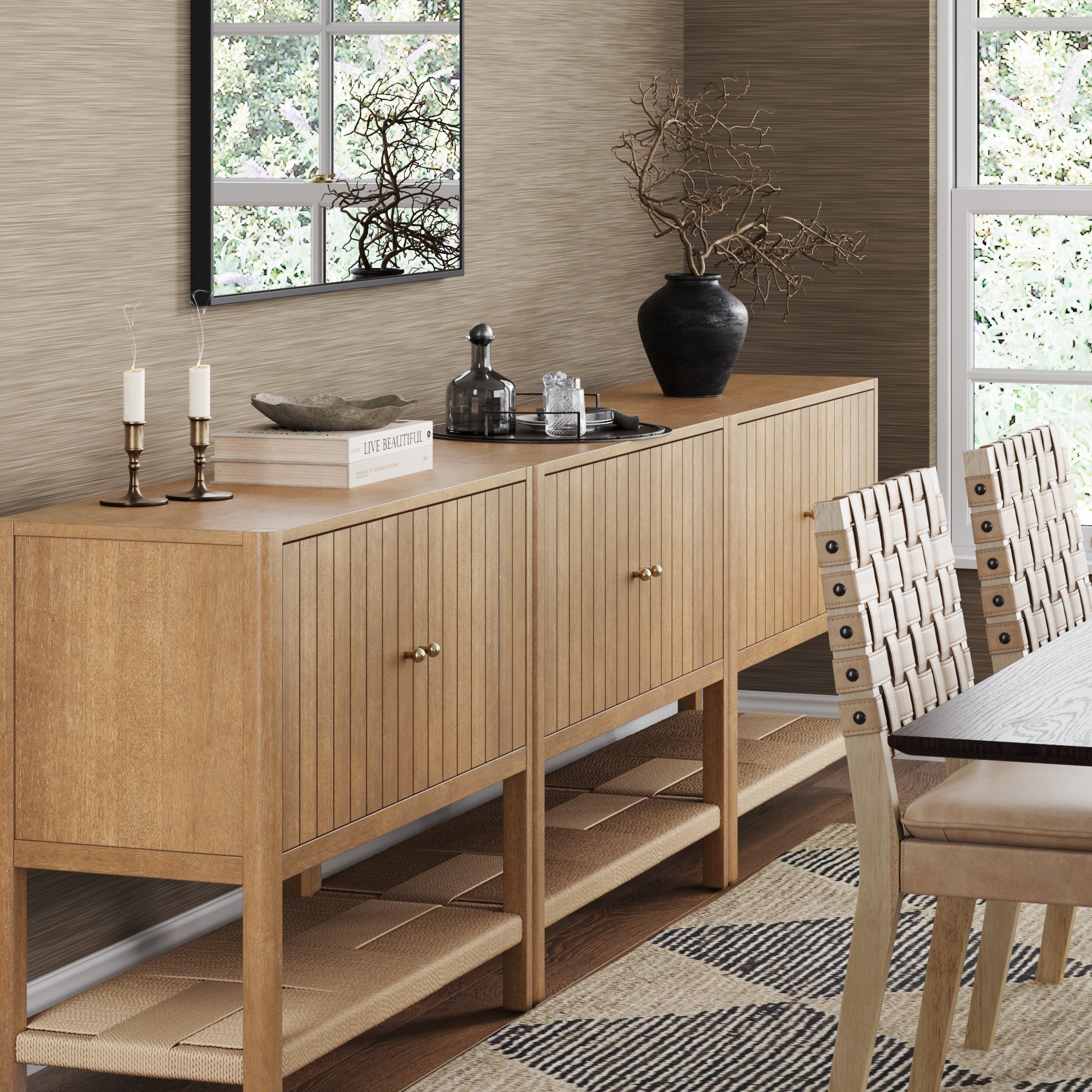 A modern dining room features the Nathan James Grooved Door TV Stand or Storage Cabinet in light brown (Set of 3) with decor like candles, books, glass bottles, and a vase with branches. A large mirror hangs above and woven chairs sit by the dining table.