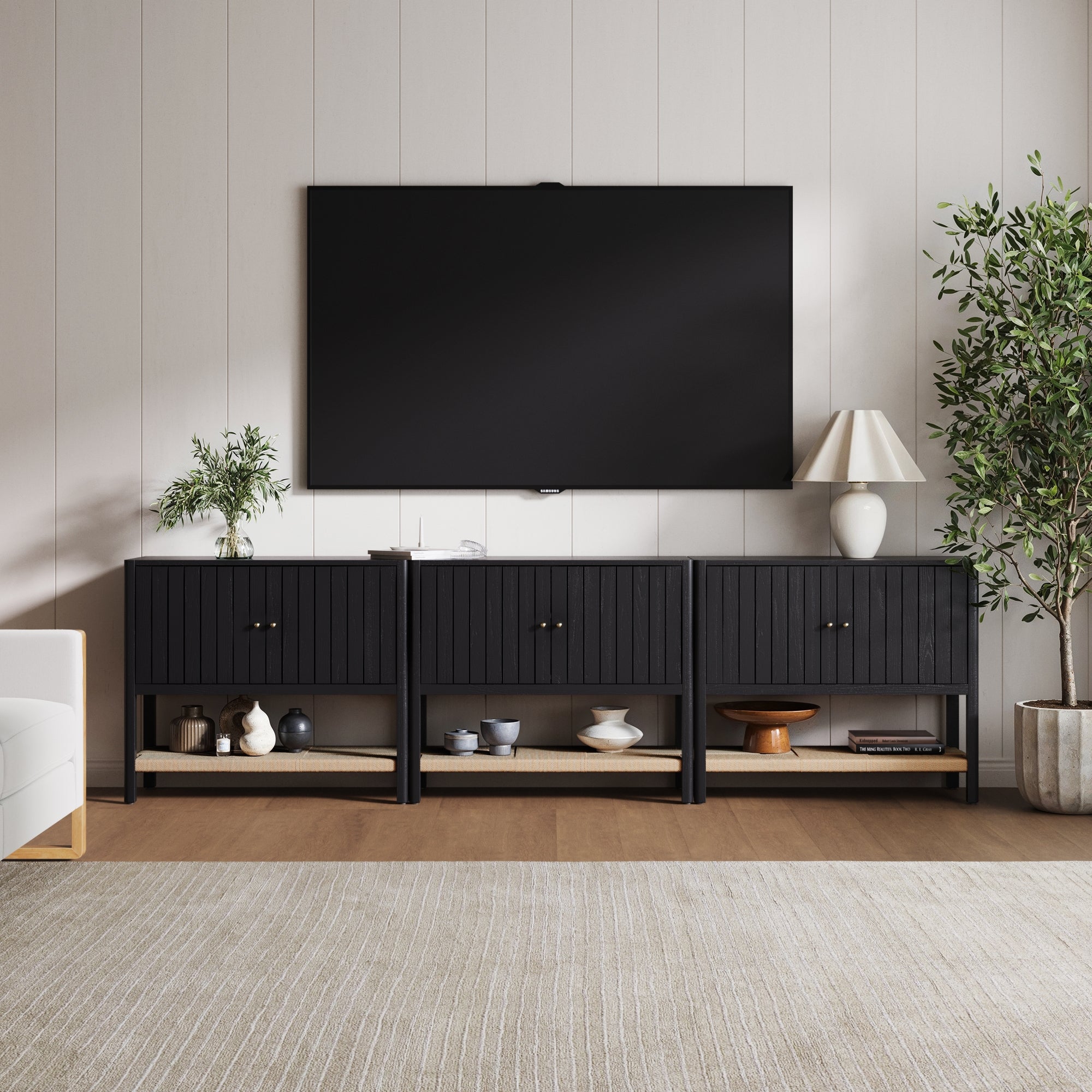 A modern living room with a wall-mounted TV above the Nathan James Grooved Door TV Stand or Storage Cabinet (Set of 3) in Black Oak, styled with plants, a lamp, and books on a light wood floor with a beige area rug.