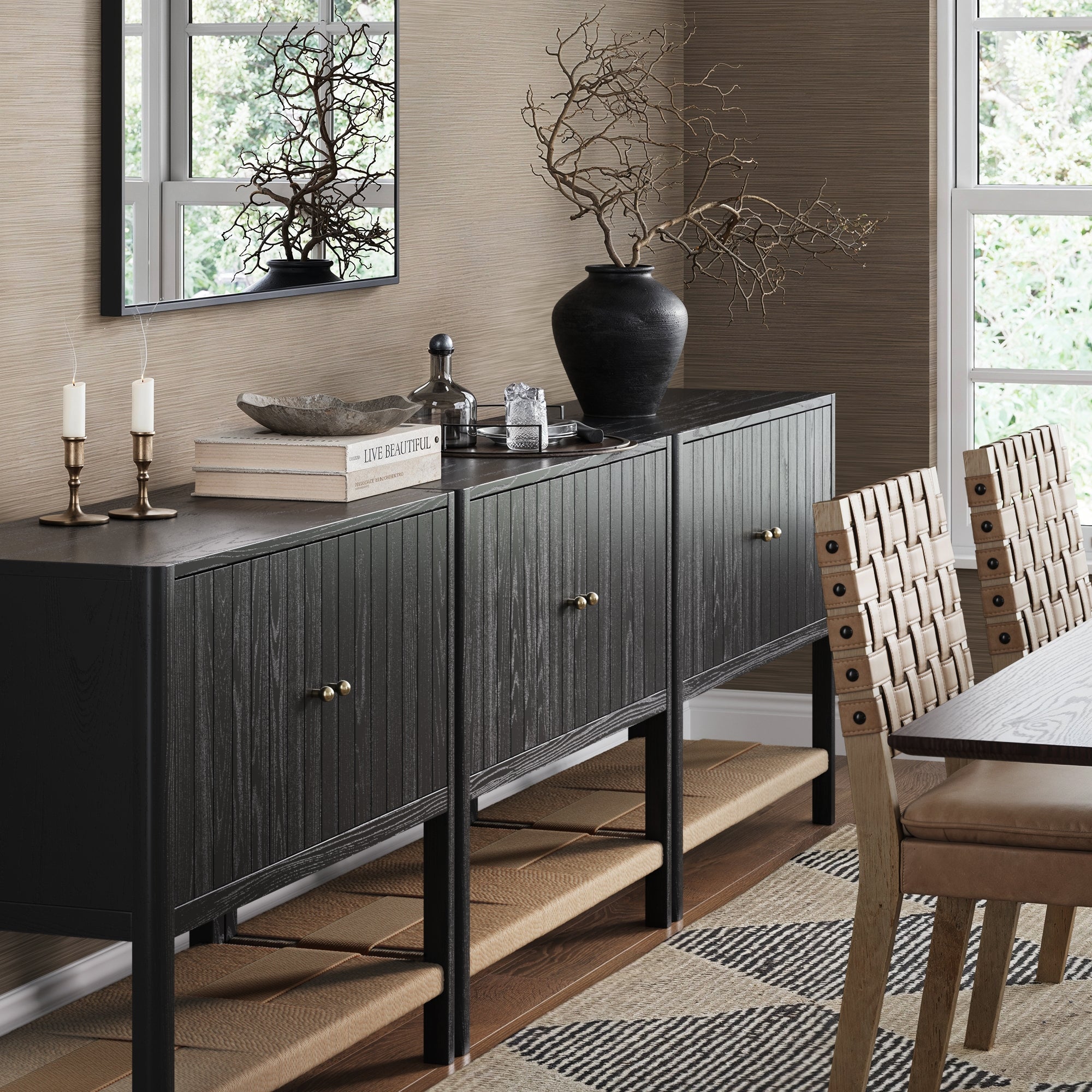 A modern dining room features the Nathan James Grooved Door TV Stand or Storage Cabinet in Black Oak (Set of 3), a mirror, woven chairs, candle holders, books, and a black vase with branches. Large windows brighten the space over a patterned rug.