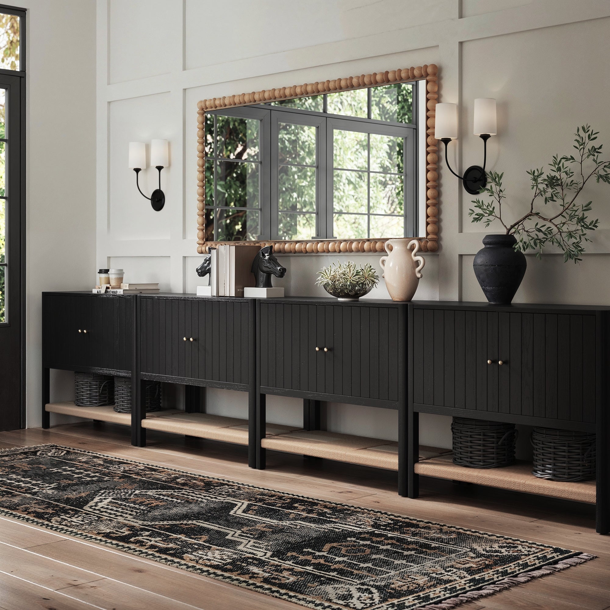 A modern hallway showcases the Nathan James Grooved Door Credenza TV Console in Black Oak (Set of 4) with solid wood legs, decorative vases, plants, baskets, a large wood-framed mirror above, wall sconces on each side, and a patterned rug on the wooden floor.