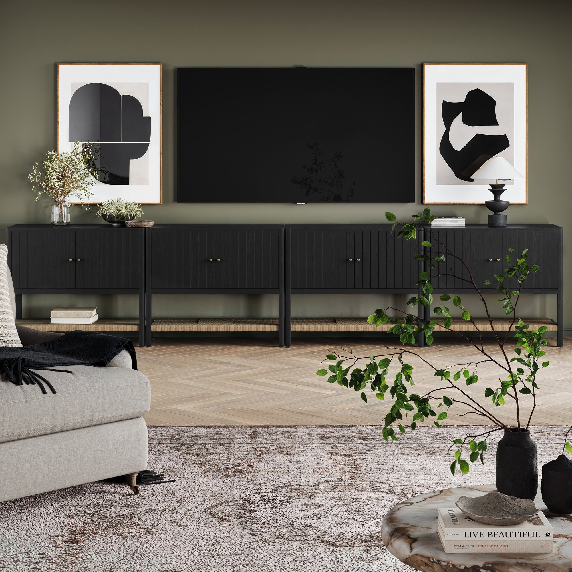 A modern living room with a Grooved Door Credenza TV Console in Black Oak by Nathan James, large TV, abstract wall art, potted plants, beige sofa with striped throw, and a round coffee table with a vase of green branches.