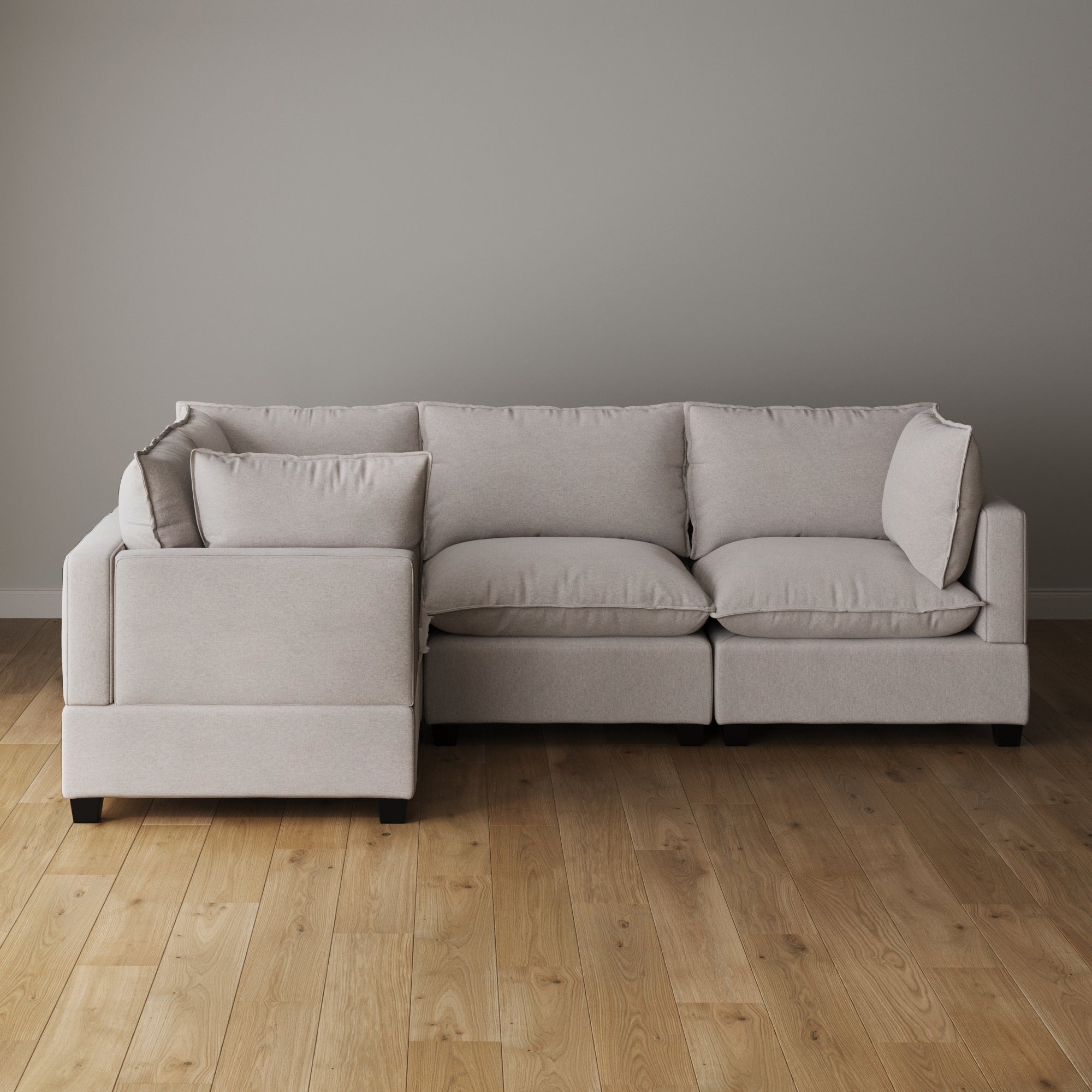 A light beige L-shaped sectional sofa with plush cushions and a low backrest sits on a wooden floor against a plain light gray wall.