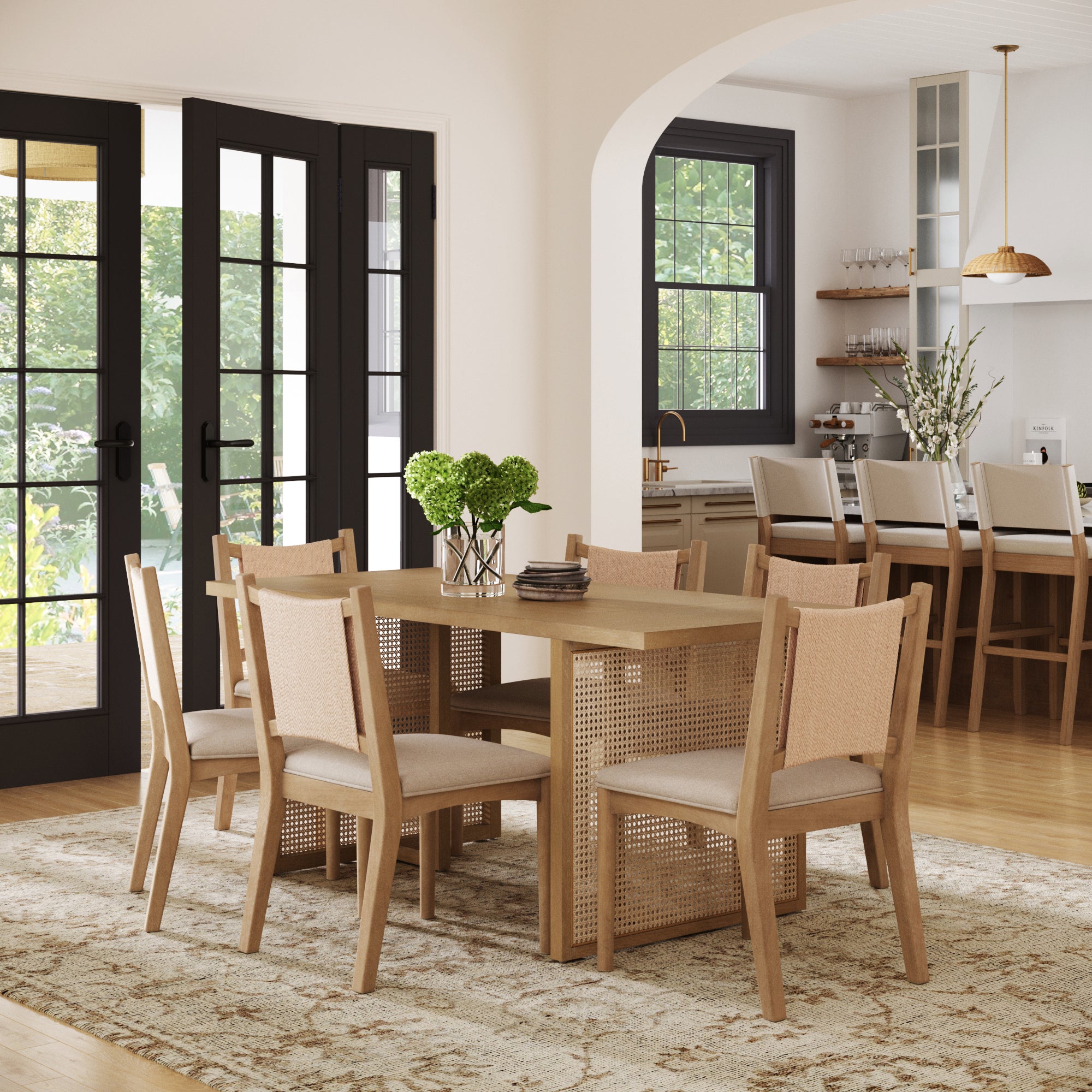 A dining room featuring the Nathan James Rattan Boho Dining Table and a set of 6 matching Seagrass Chairs, complemented by a vase of green hydrangeas and large black-framed glass doors. A kitchen with extra seating is visible in the background.