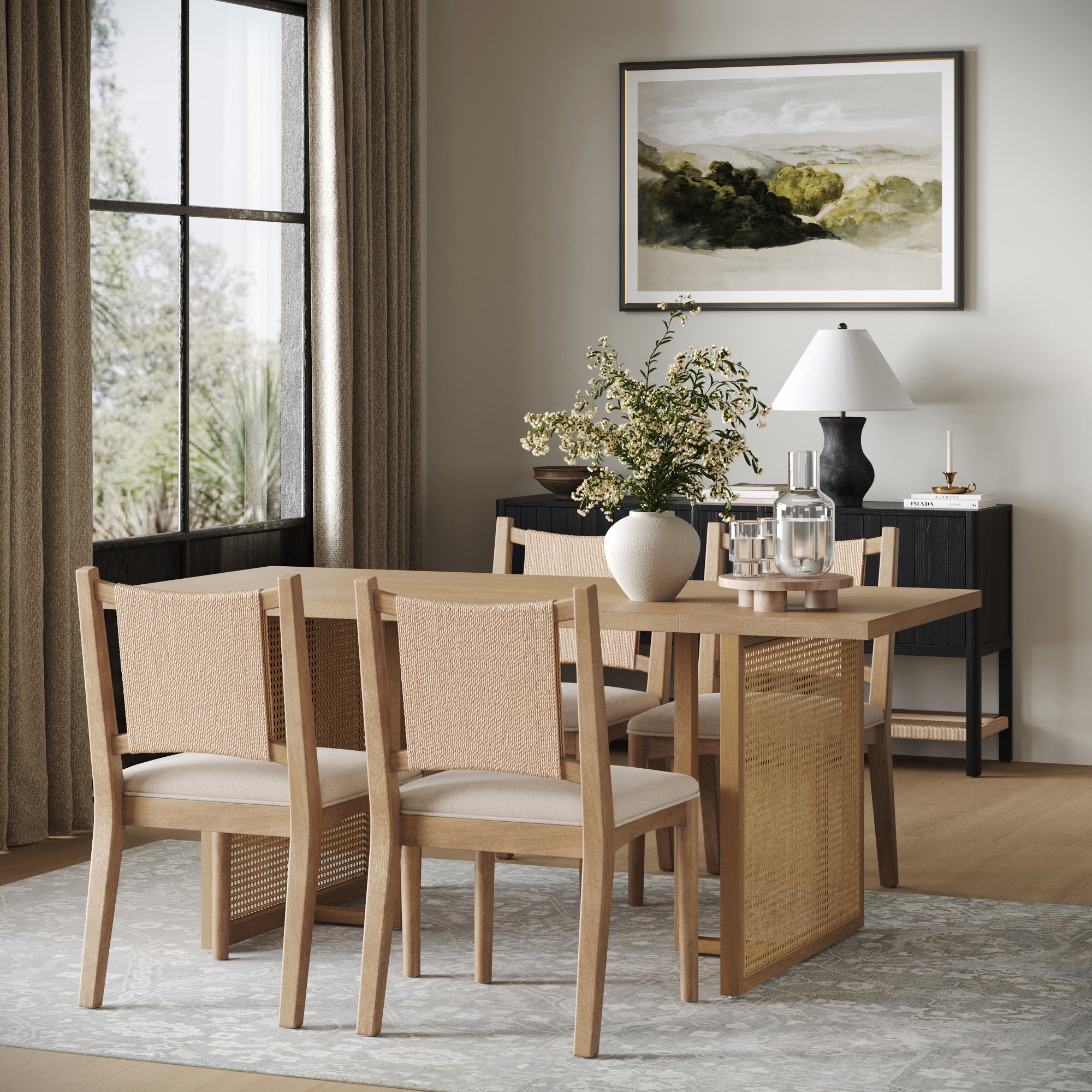 A modern dining room features the Nathan James Rattan Boho Dining Table with Seagrass Chairs Set of 4, a light rug, a black sideboard with lamp and vase, a landscape painting, and large windows that fill the space with natural light.