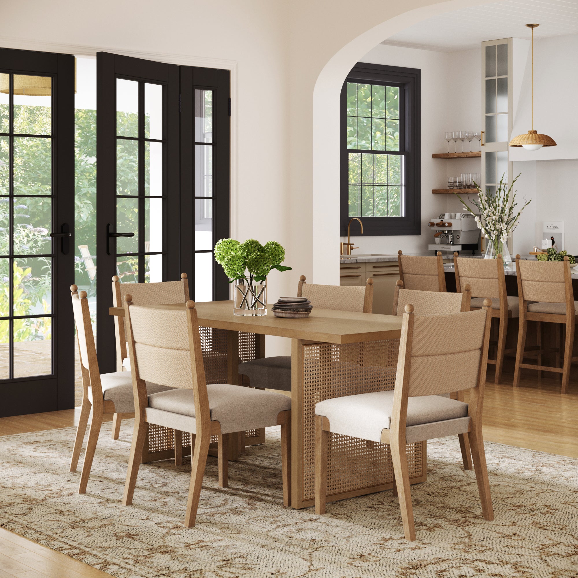 A modern dining room showcases the Nathan James Rattan Dining Table with Flax Upholstered Chairs Set of 6, complemented by a rug and green hydrangea vase. Large windows brighten the space, with an open archway leading to a stylish kitchen.