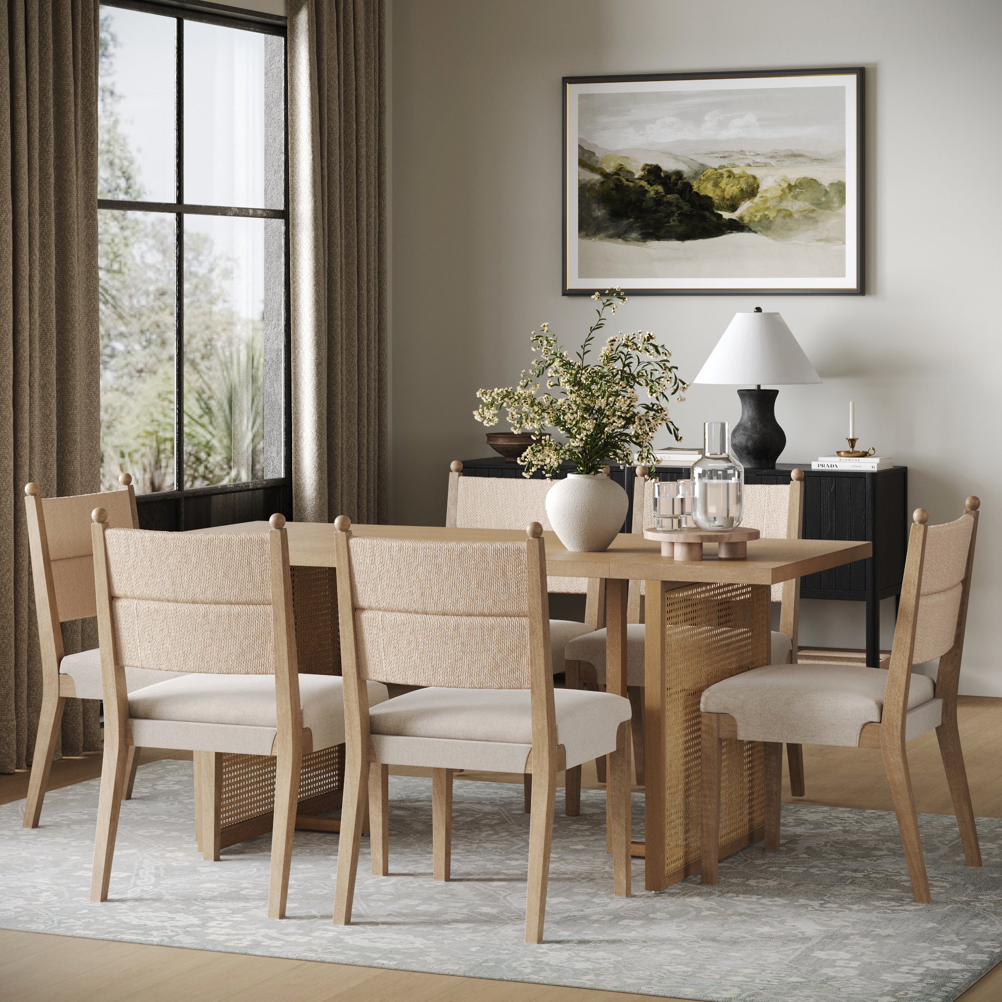 A modern dining room with the Nathan James Rattan Dining Table and six flax upholstered chairs, a sideboard topped with a white lamp and decor, framed landscape art, and a large window with curtains letting in natural light.