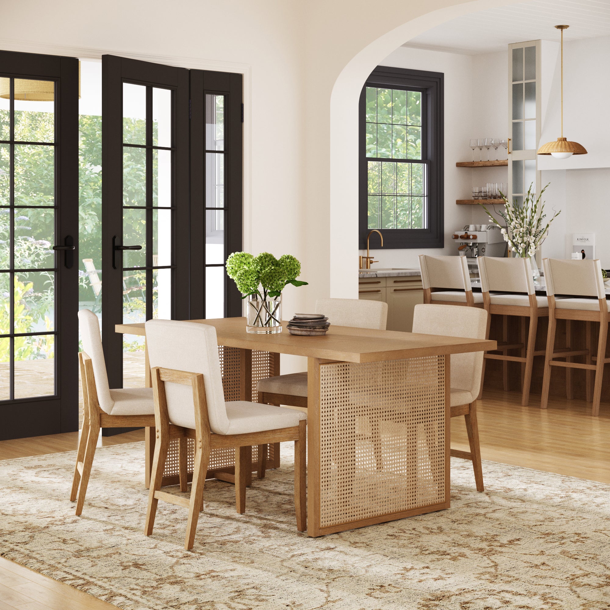 A modern dining room features the Nathan James Rattan Boho Dining Set with four upholstered chairs. Large black-framed windows fill the space with light, while a kitchen with barstools and pendant lighting sits in the background.