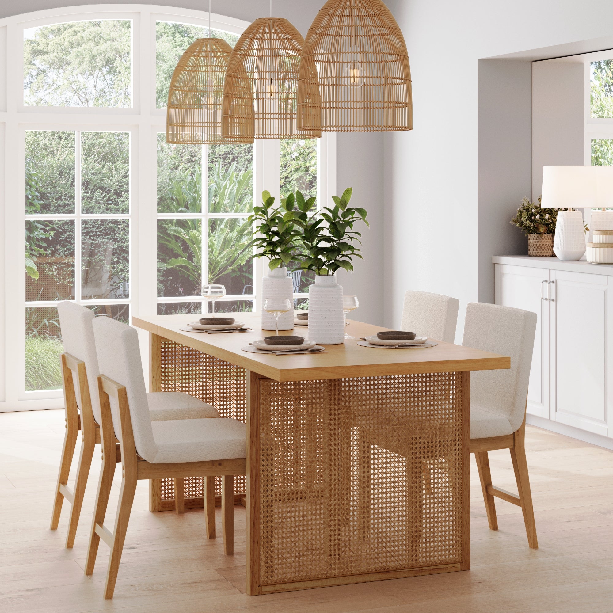 A modern dining room features the Nathan James Bohemian Natural Wood & Rattan Dining Table, six cushioned chairs, woven rattan pendant lights, white vases with greenery, and large windows that flood the airy space with natural light.