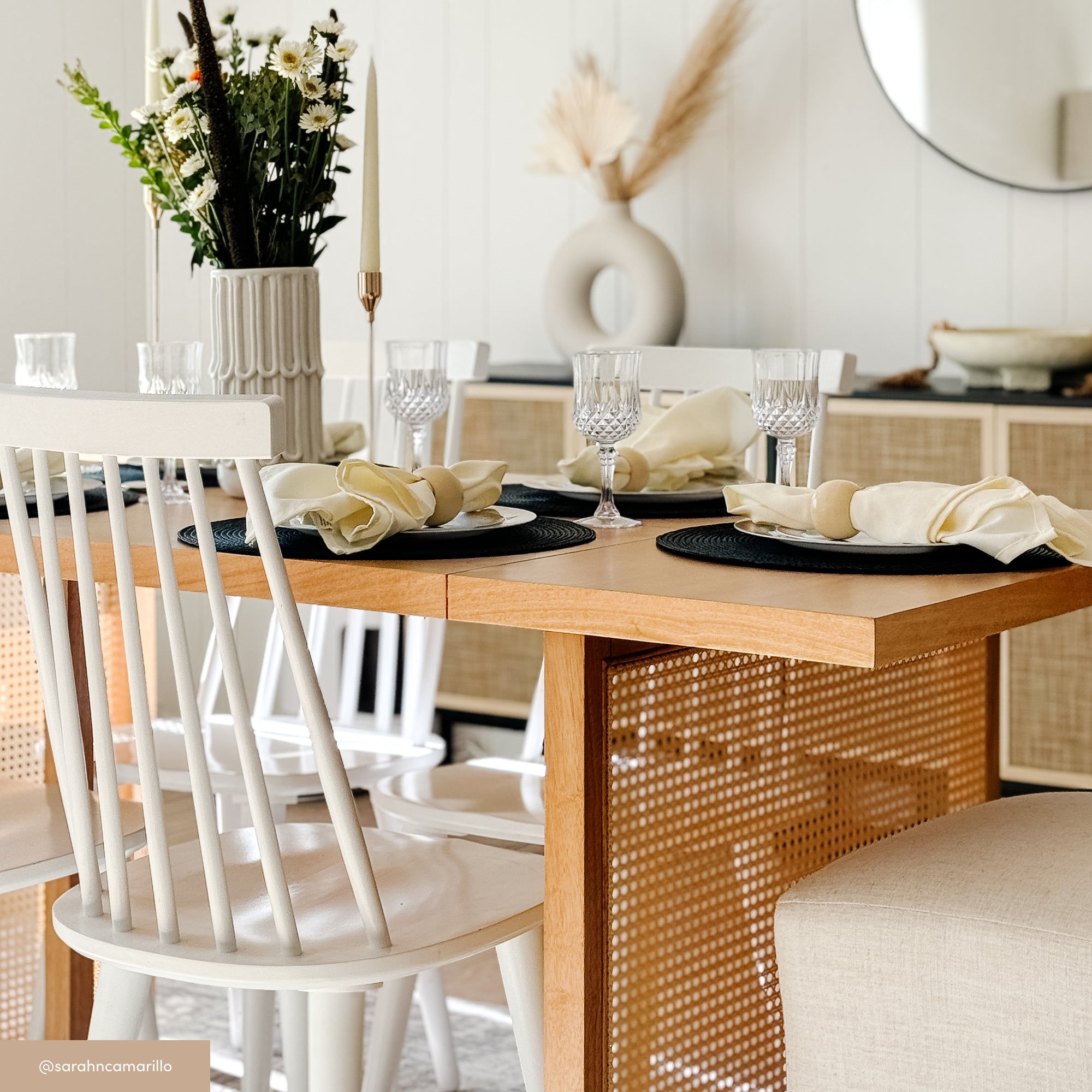 A modern dining table set with four white chairs, beige napkins, glassware, and a floral centerpiece. The background features light wood furniture, pampas grass decor, and a round mirror on a white wall.