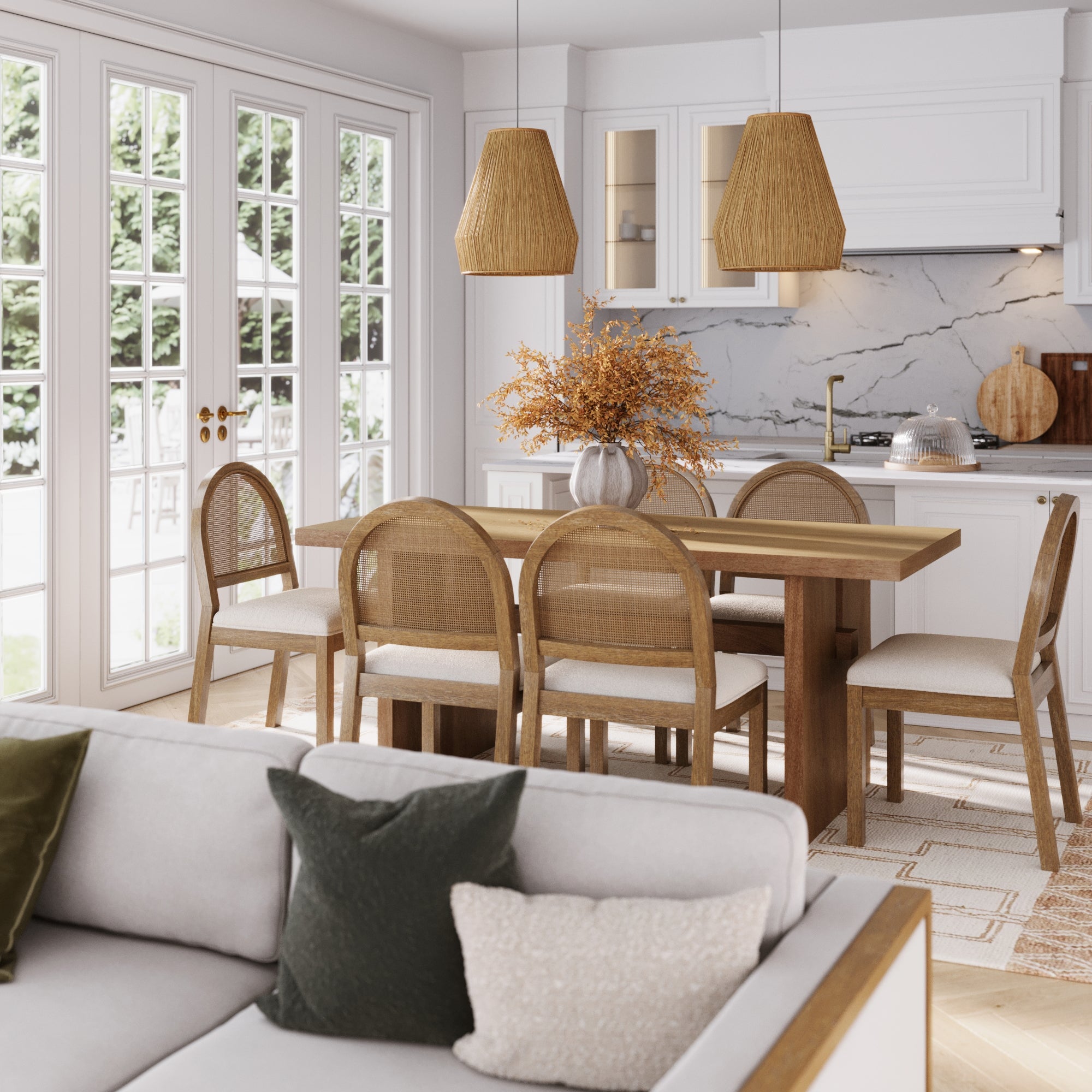 A modern dining area showcases the Nathan James Bohemian Cane Dining Set with six chairs, a vase of dried flowers, pendant lights, and a kitchen in the background. Large windows fill the space with light, while a cushioned sofa sits in the foreground.
