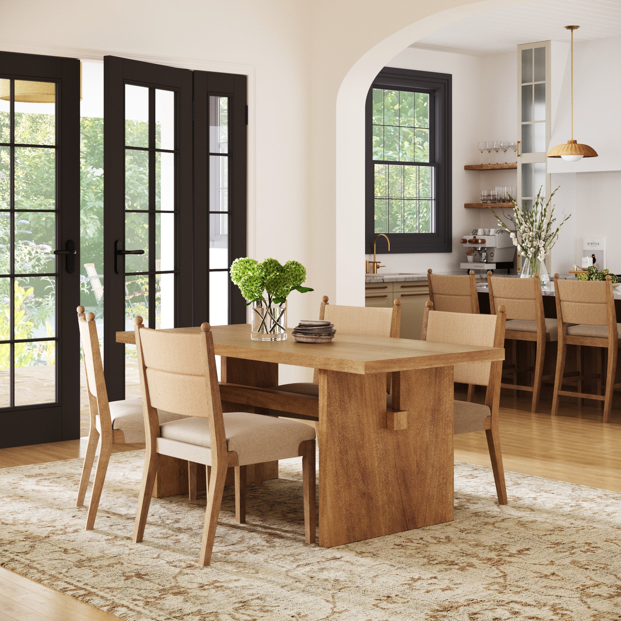 A modern dining room featuring the Nathan James Boho Solid Wood Dining Table Set for 4 in light brown, paired with a beige rug, black-framed glass doors, and an open view of a coordinated kitchen with a vase of flowers on the counter.