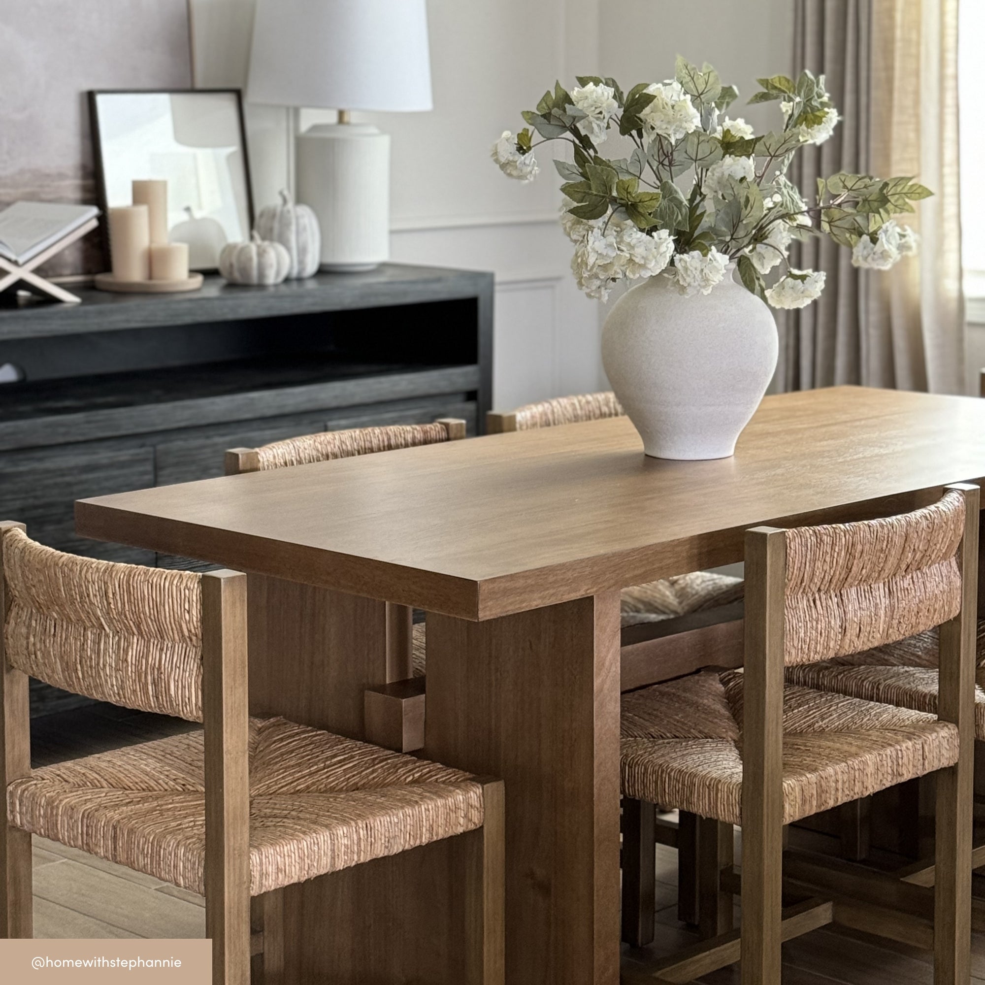 A wooden dining table with four woven chairs is topped with a white vase holding white flowers. In the background, a console table displays candles, books, and framed artwork. The room has neutral tones and natural light.