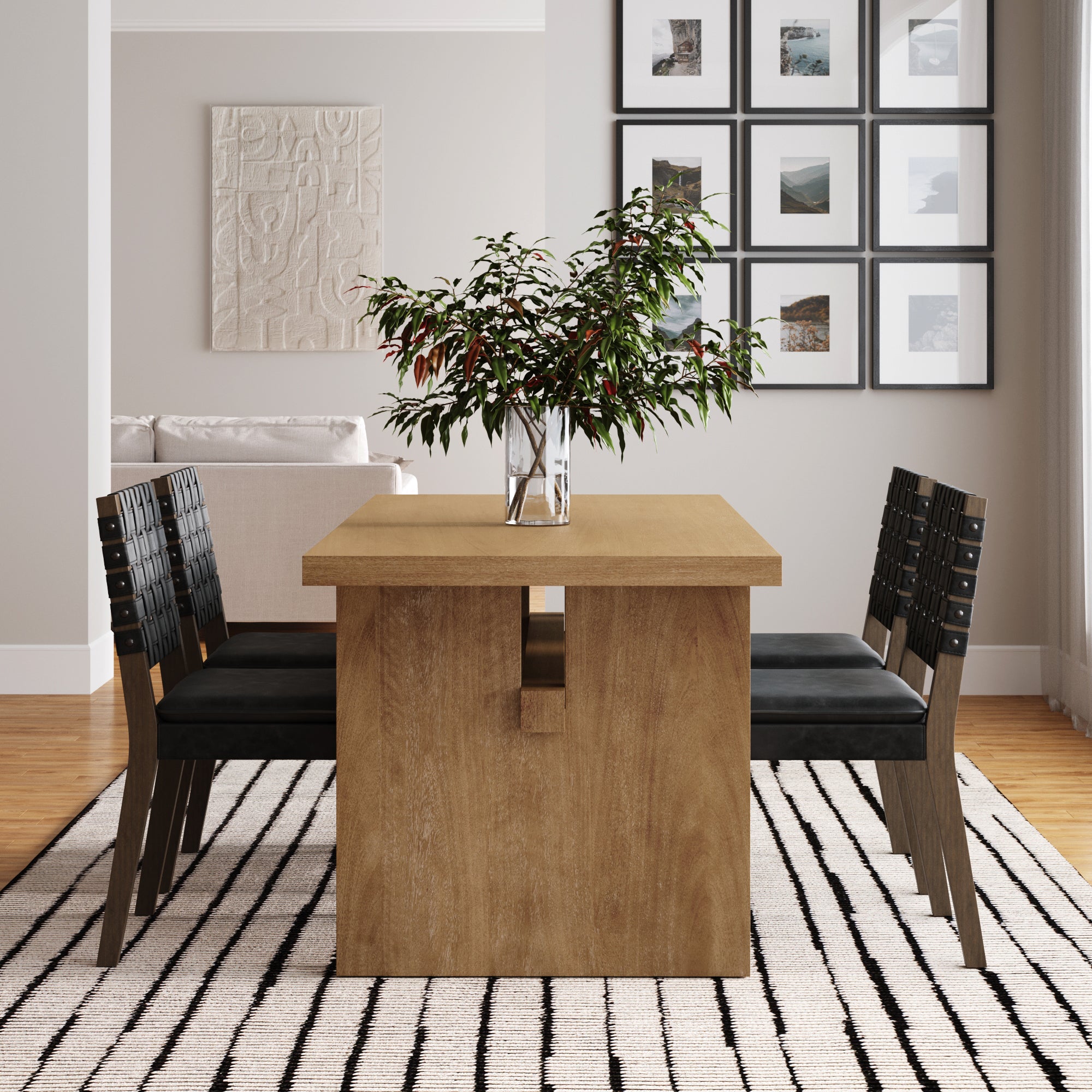 A modern dining room with a wooden table, four black chairs, a vase with green leafy branches, a striped rug, and framed art on the wall. A beige couch and textured artwork are visible in the background.