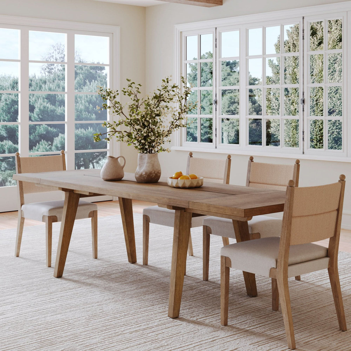 Nathan James Farmhouse Extendable Wood Dining Table with Leaf Extensions in light brown, rectangular with four upholstered chairs, set with a vase of branches and a bowl of lemons.