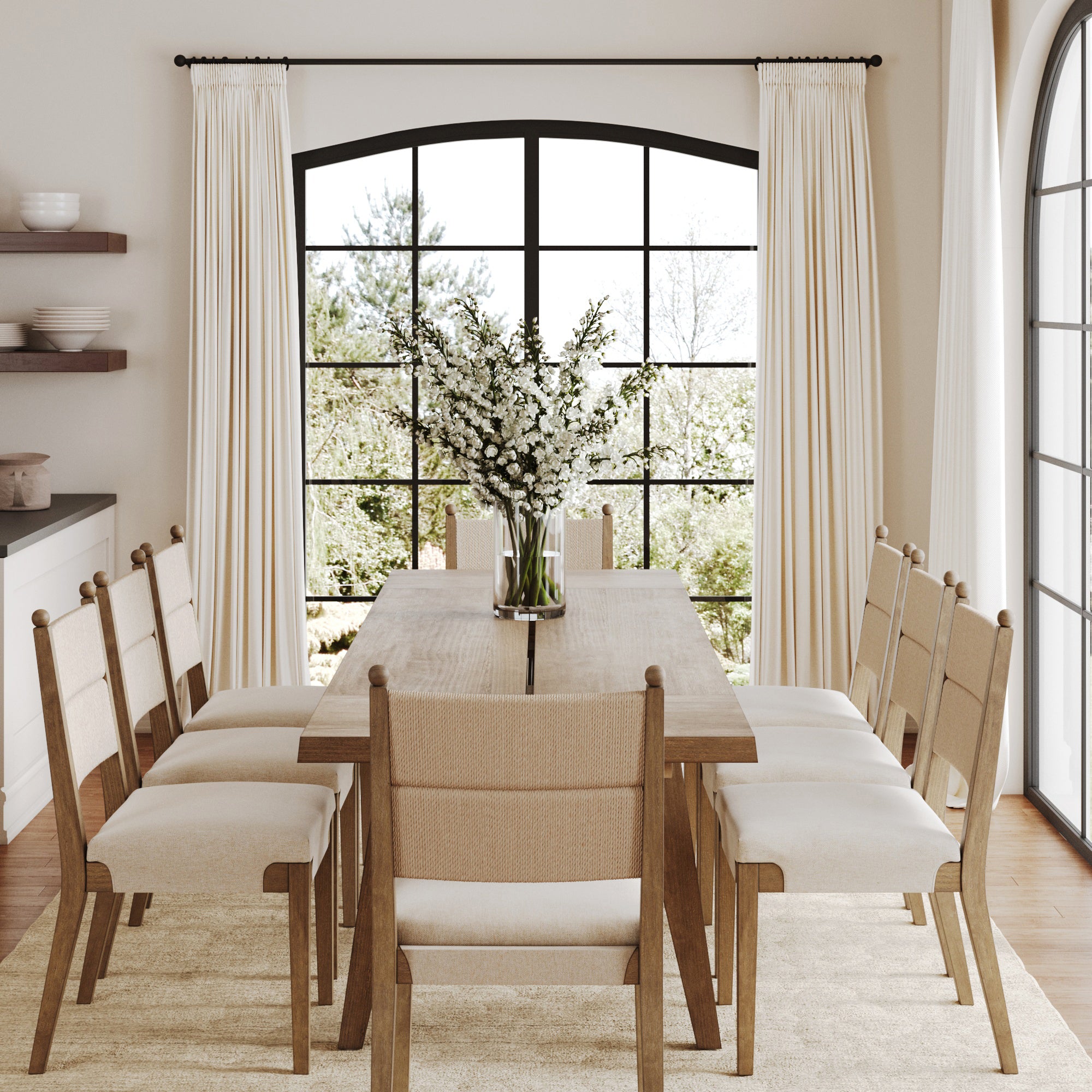 A bright dining room with a Nathan James Farmhouse Wooden Dining Table with Leaf Extension in light brown, eight beige cushioned chairs, a vase of white flowers, cream curtains, black-framed windows, and views of greenery.