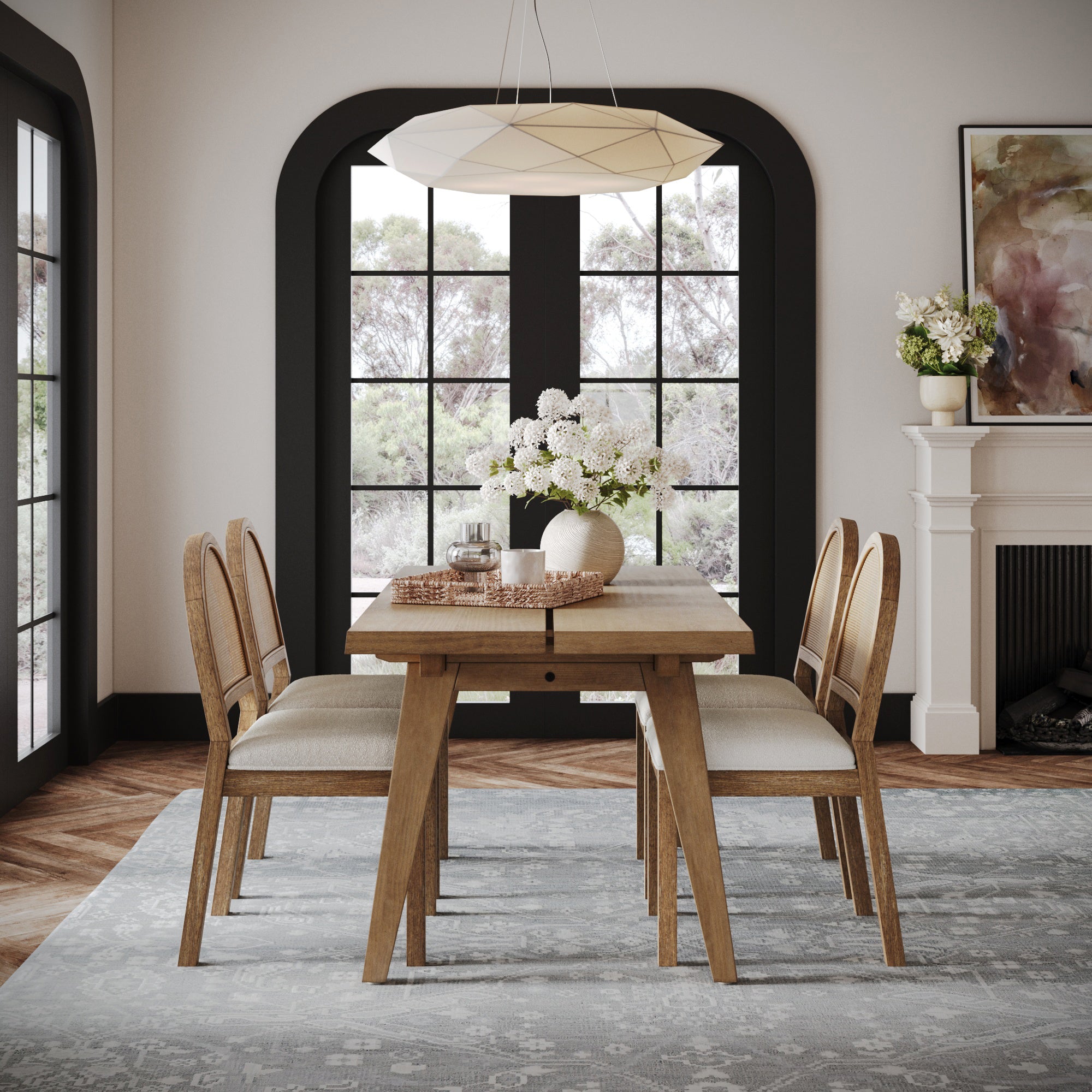 A cozy dining room features the Nathan James Modern Farmhouse Extendable Dining Table in light brown, beige cushioned chairs for four, a white flower centerpiece, arched windows, a modern pendant light, and a white fireplace with decor.