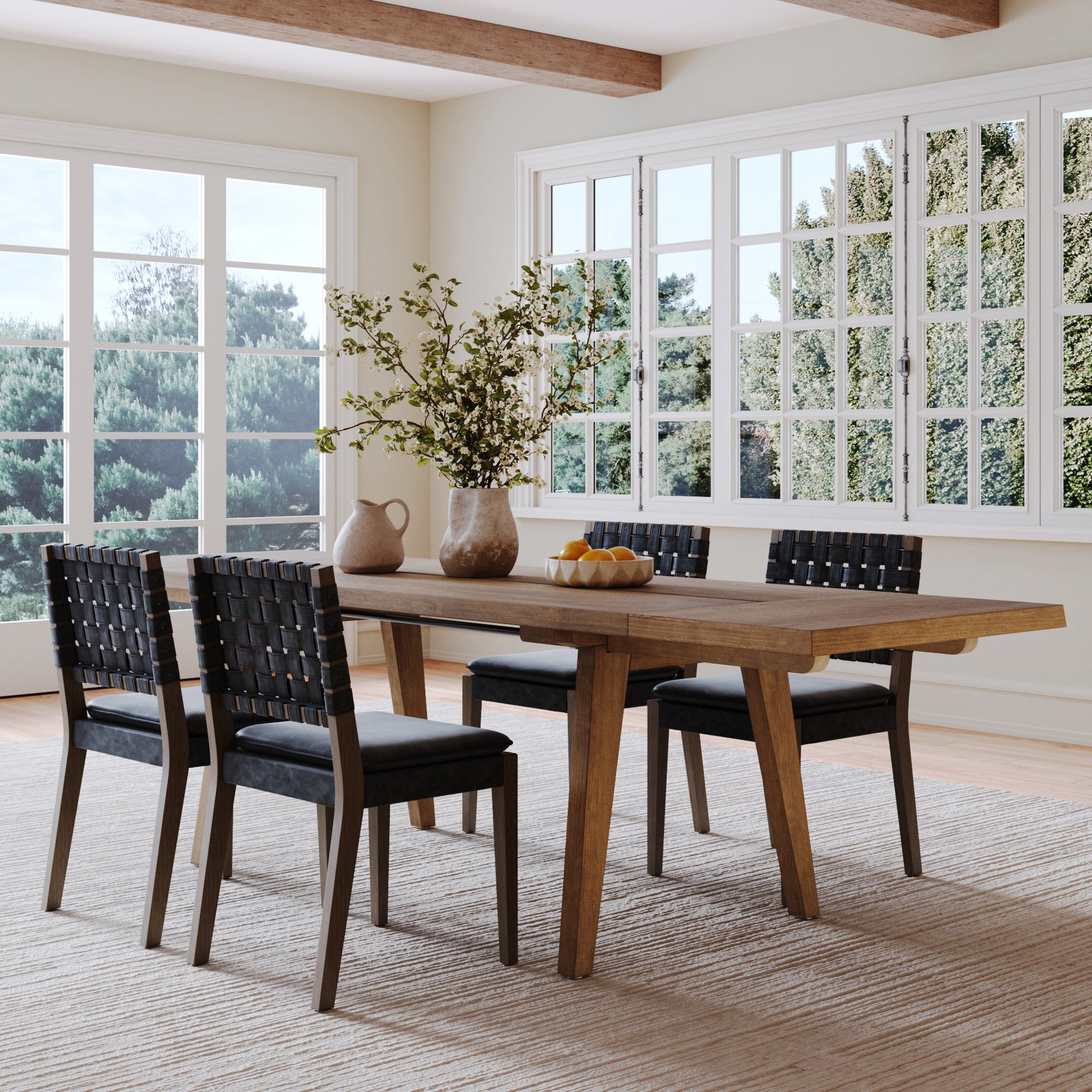 A modern dining room features the Nathan James Extendable Dining Set with four black dining chairs, a vase with green branches, a white pitcher, and a bowl of oranges. Large windows provide scenic tree views.