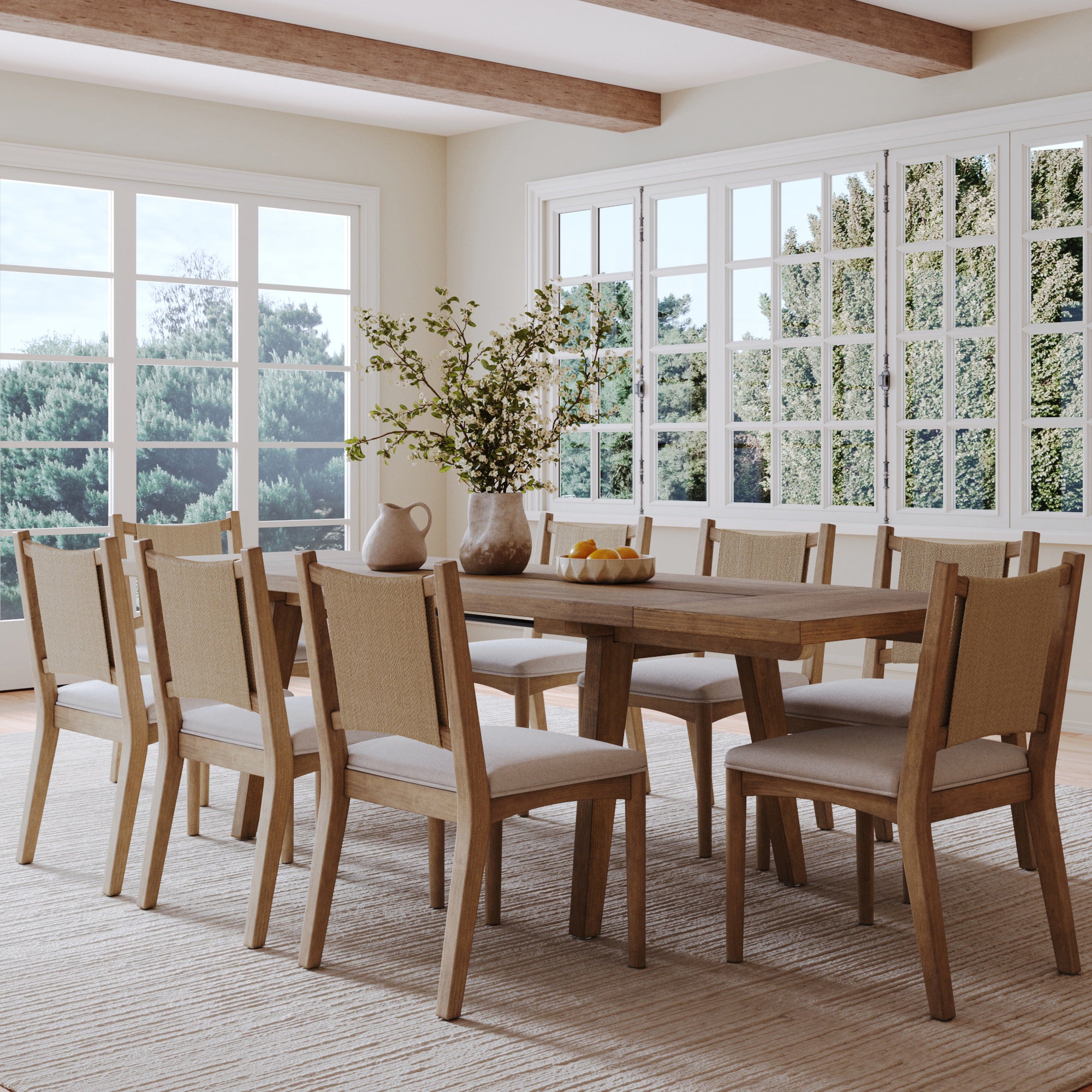 A bright dining room with large windows features the Nathan James Bohemian Wooden Dining Table Set, seating eight, paired with beige chairs and styled with a vase of branches, a bowl of lemons, and a ceramic pitcher.